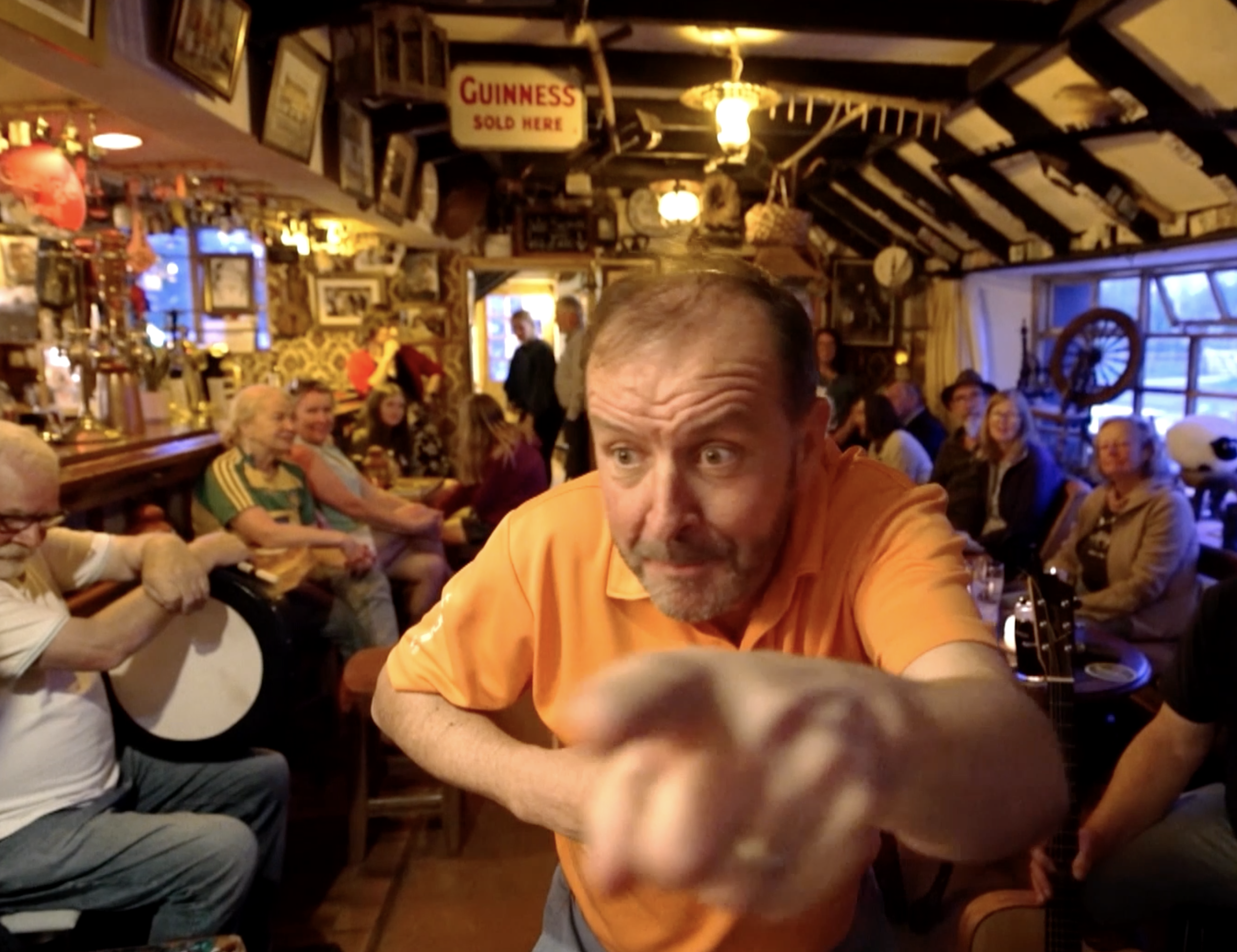 A crowded pub with a man in an orange shirt reaching toward the camera. The pub has wooden decor, vintage signs, and people sitting and standing, some playing instruments and others socializing.