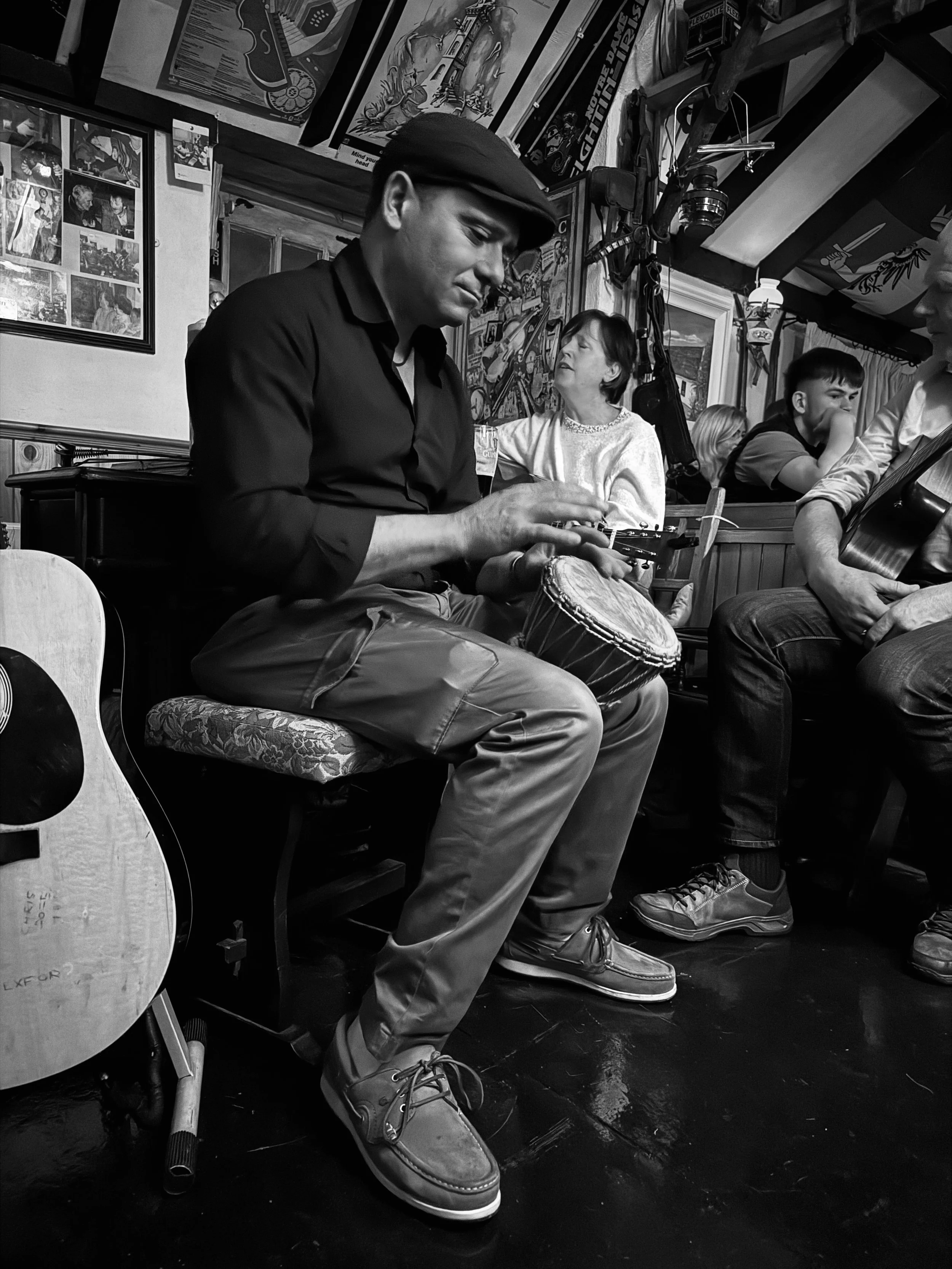 A man playing a drum in a lively, decorated restaurant or bar with other patrons, including a woman and a man with a guitar, visible in the background.