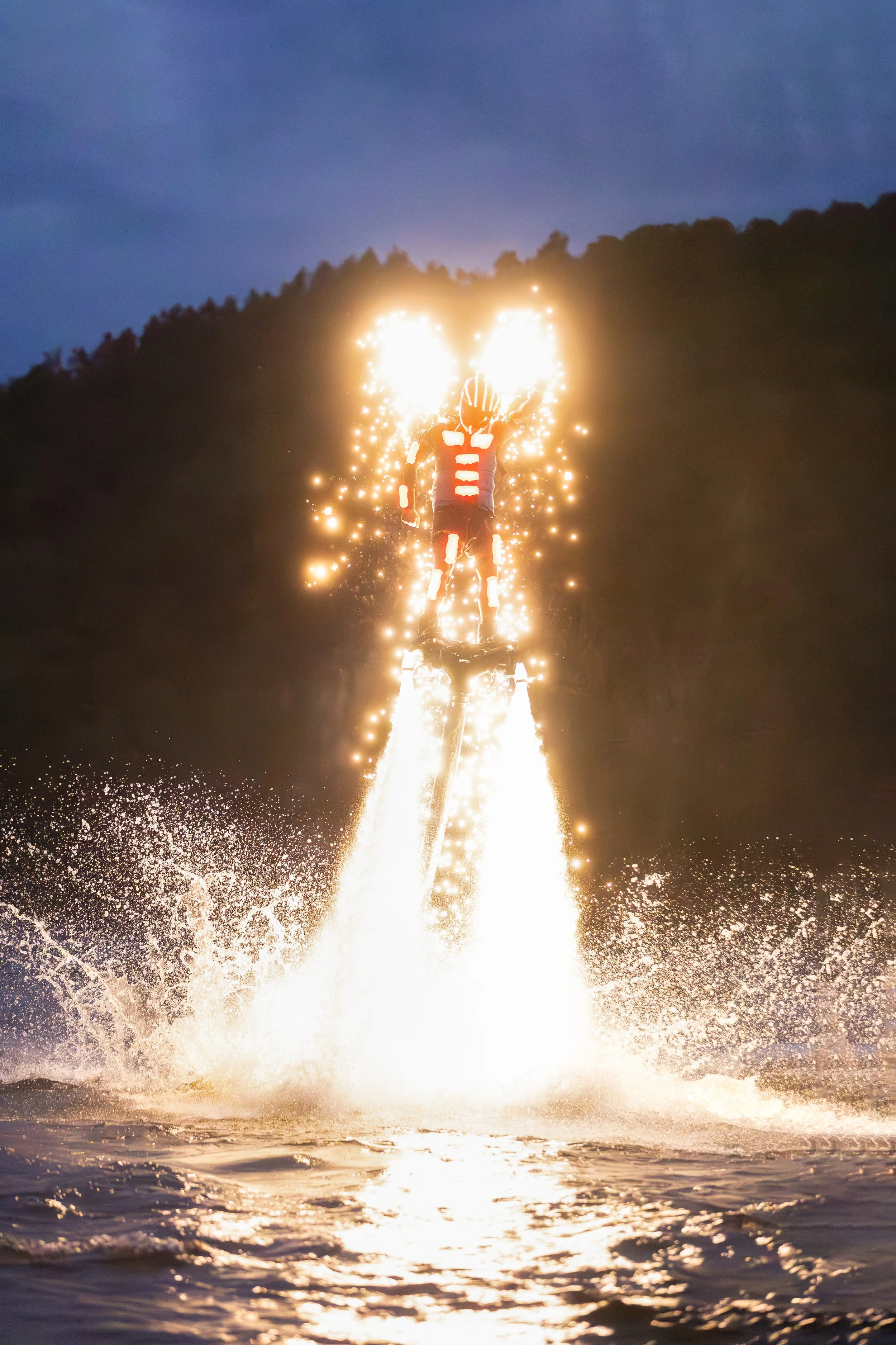 Osoba stojící na jetpacku s vodními tryskami letí nad vodou, s jiskrami a jasnými světly, během soumraku.