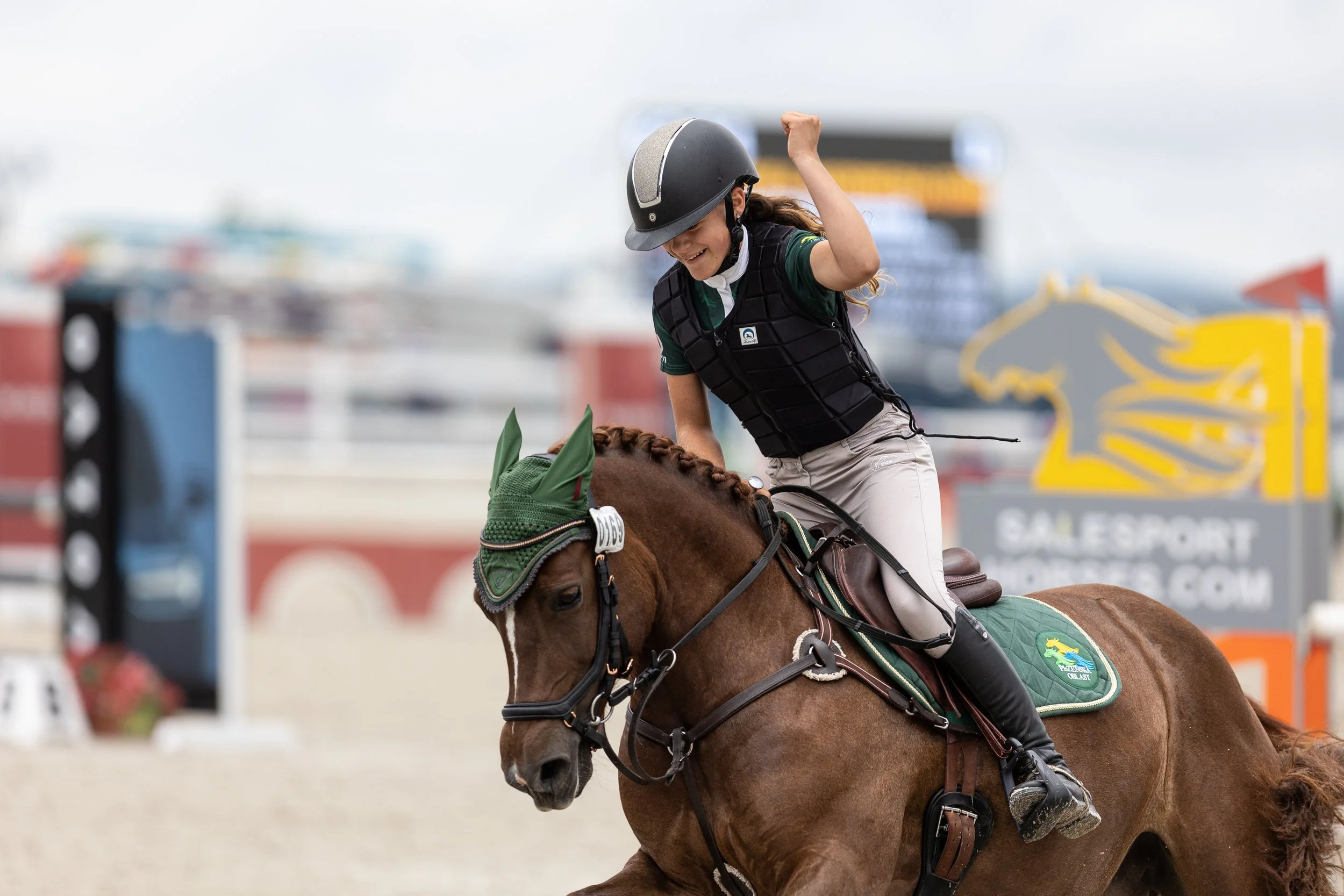 Žena na koni během parkurové soutěže, v helmě, černé ochranné vestě a béžových rajtkách, zvedá ruku na oslavu.