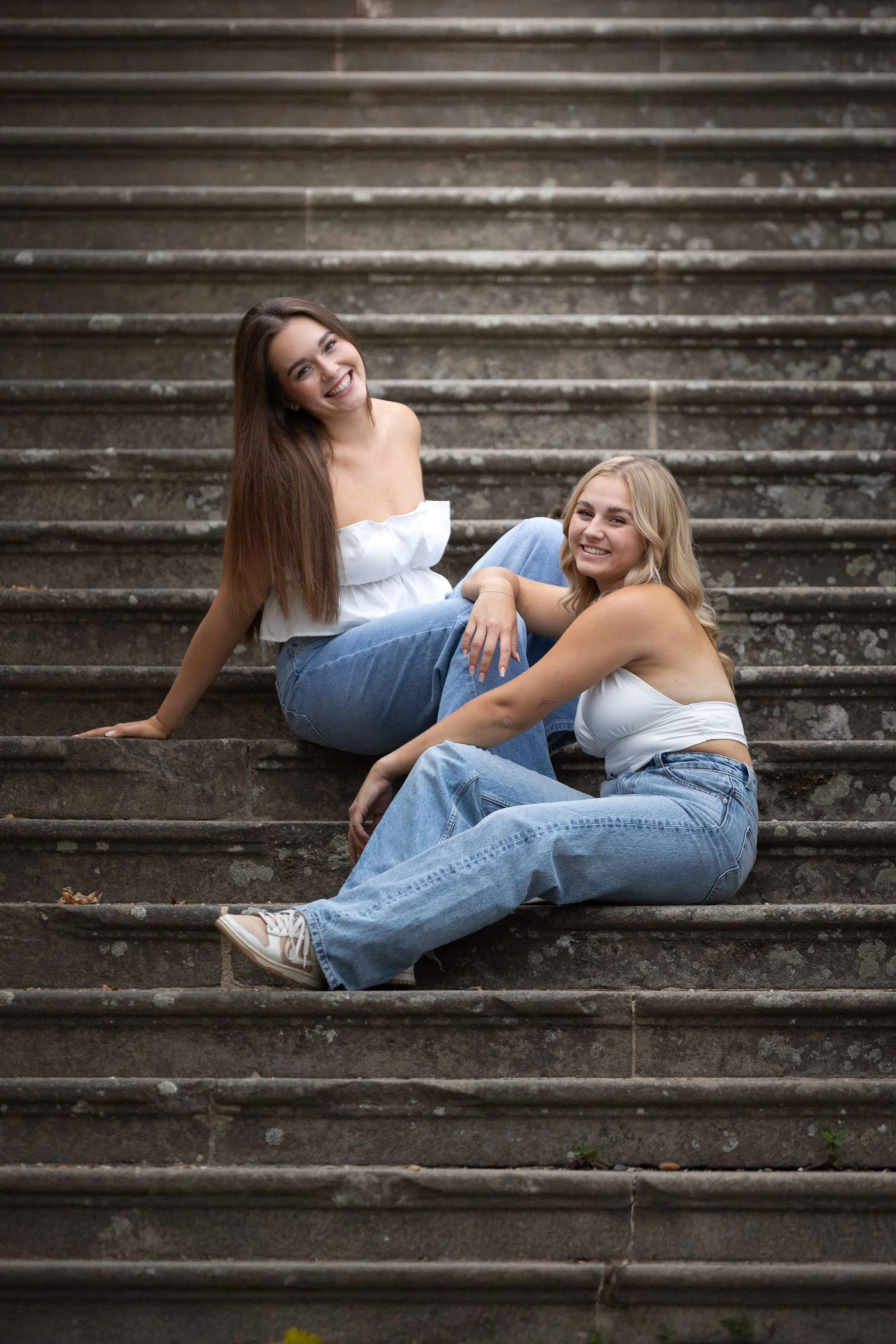 Dvě mladé ženy sedící na venkovních kamenných schodech, usmívající se a užívající si společnost té druhé.