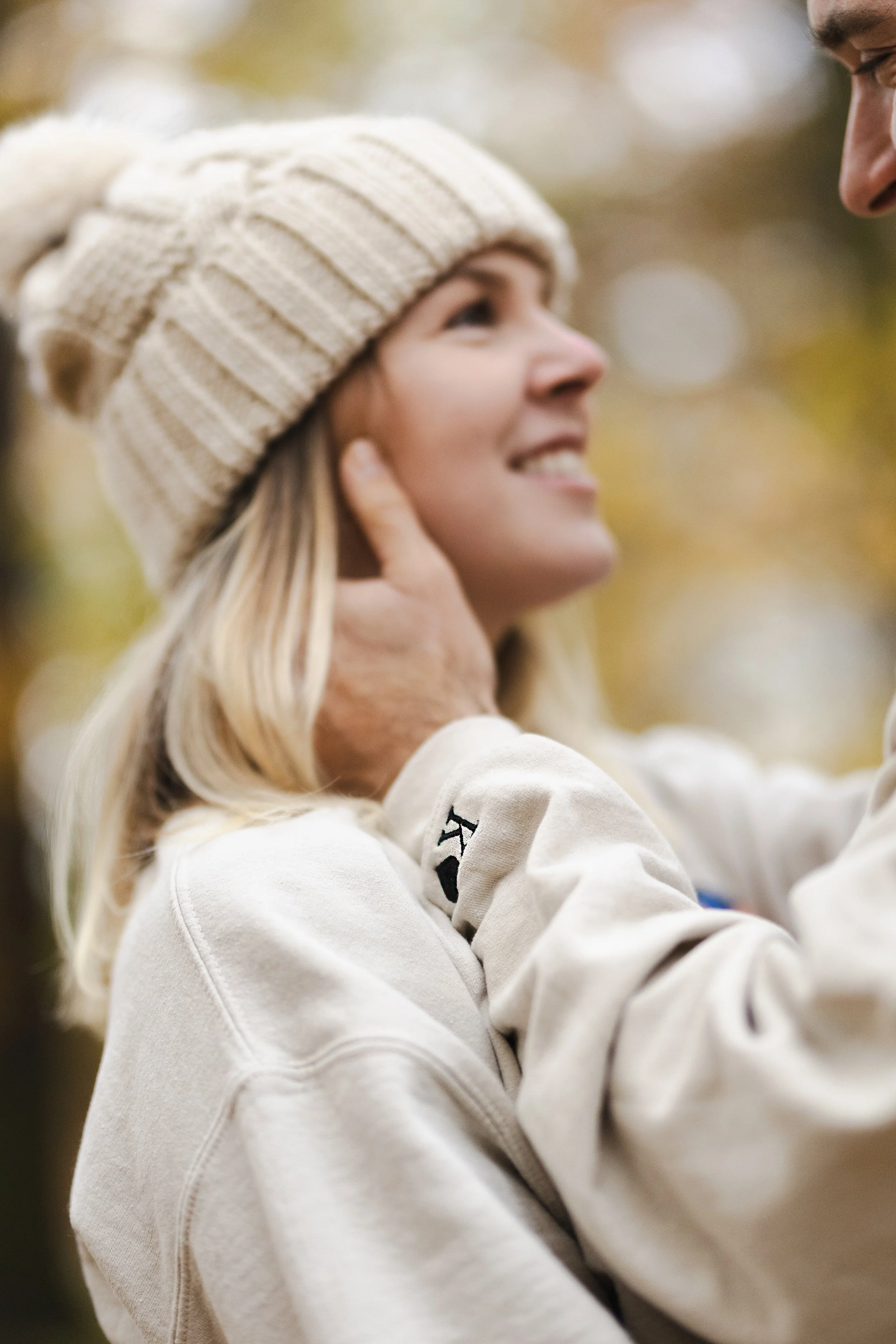 Mladá žena s blond vlasy v béžové pletené čepici a světlé mikině se usmívá, zatímco se jí někdo v bílém oblečení dotýká na krku.