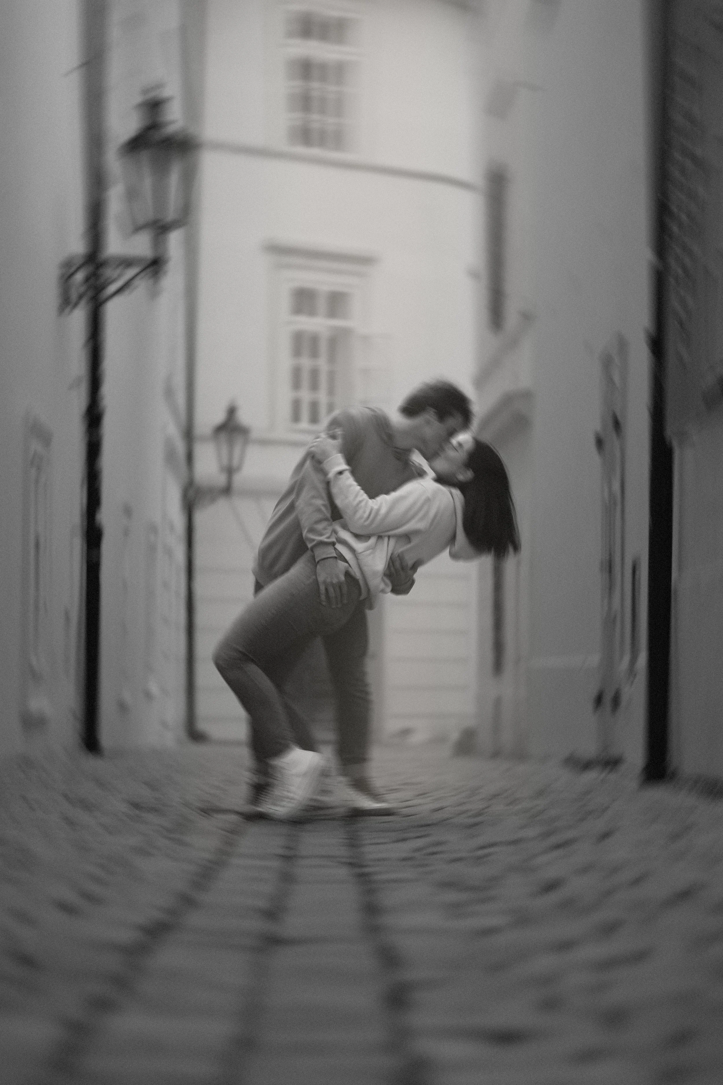 Black and white photo of a couple kissing on a narrow street, with cobblestones and old buildings around them.
