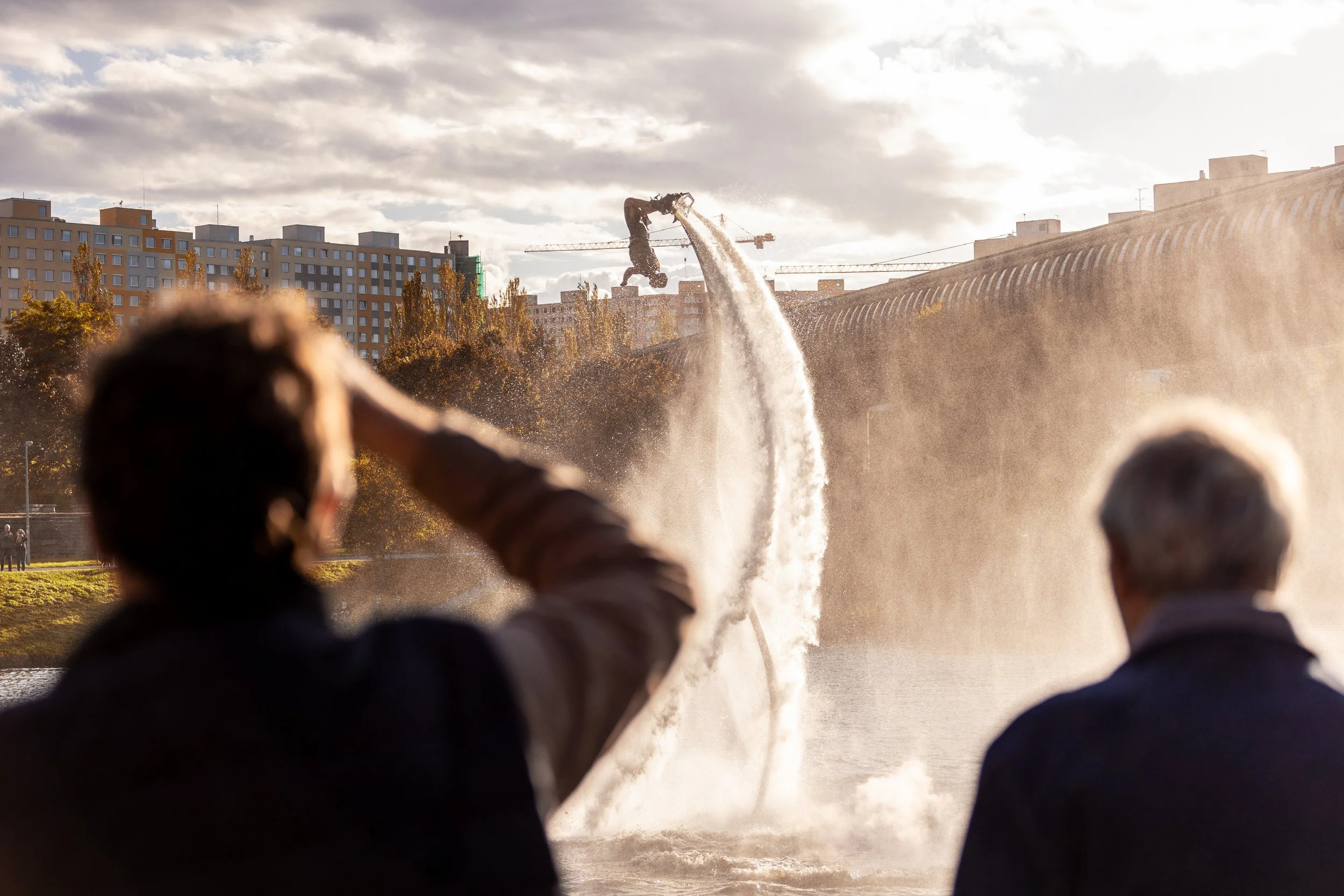 Lidé sledují fontánovou show v parku s velkým vodním gejzírem, s městskými budovami a stromy v pozadí, během pozdního odpoledne.