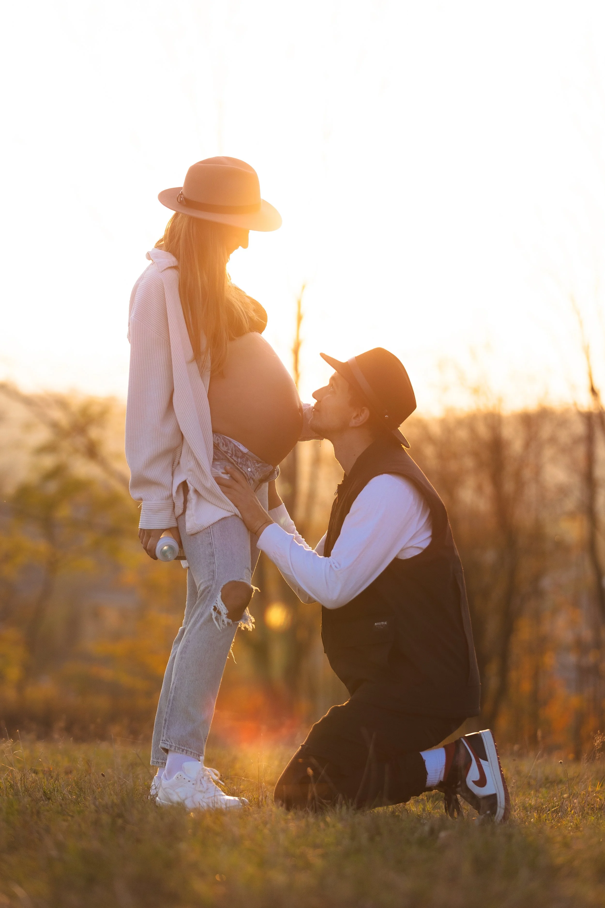 Muž s těhotnou ženu v parku při západu slunce, se stromy a teplým světlem v pozadí.