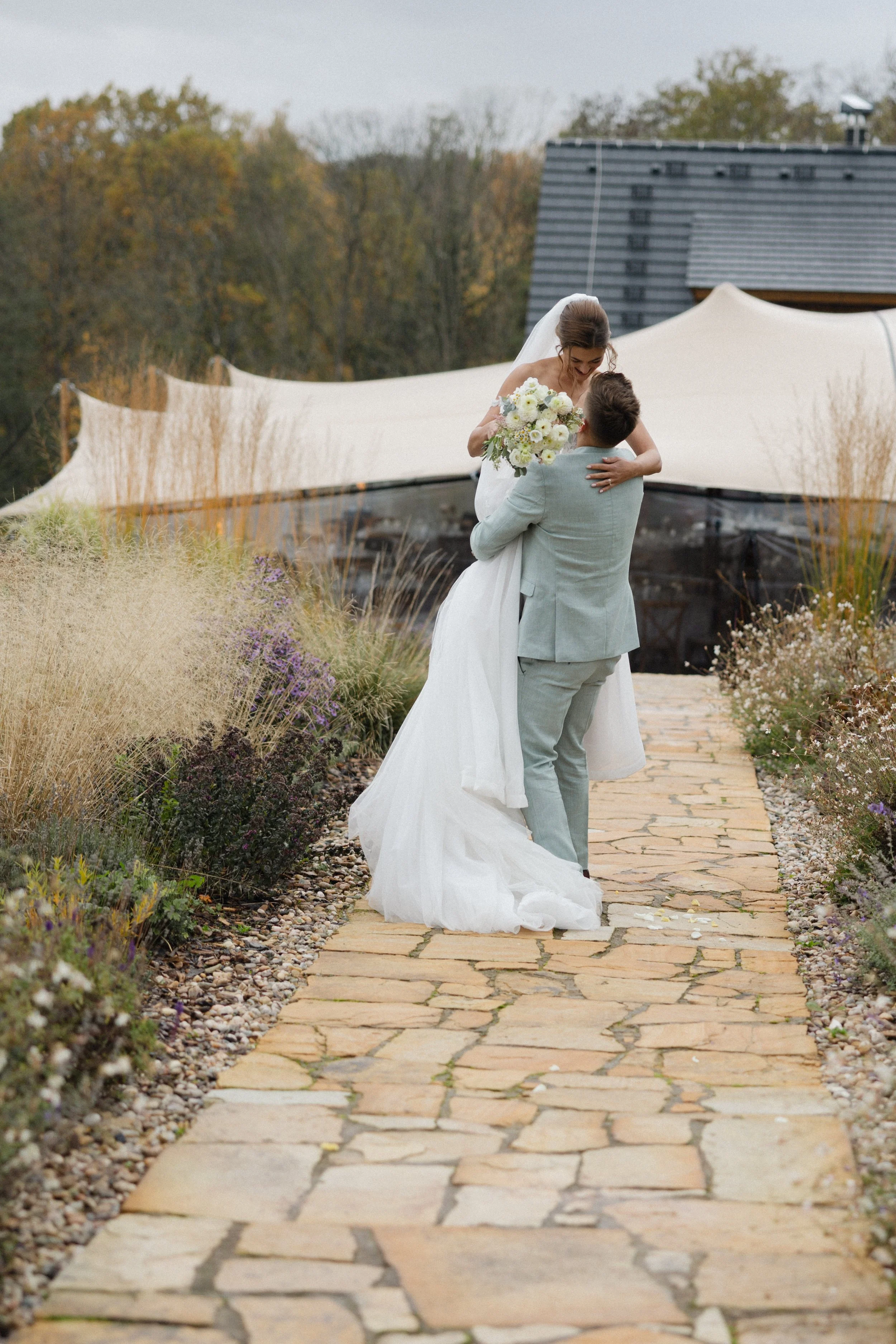 Nevěsta v bílých svatebních šatech je zvedána ženichem ve světle šedém obleku na kamenné cestě obklopené rostlinami, se stanem a stromy v pozadí.