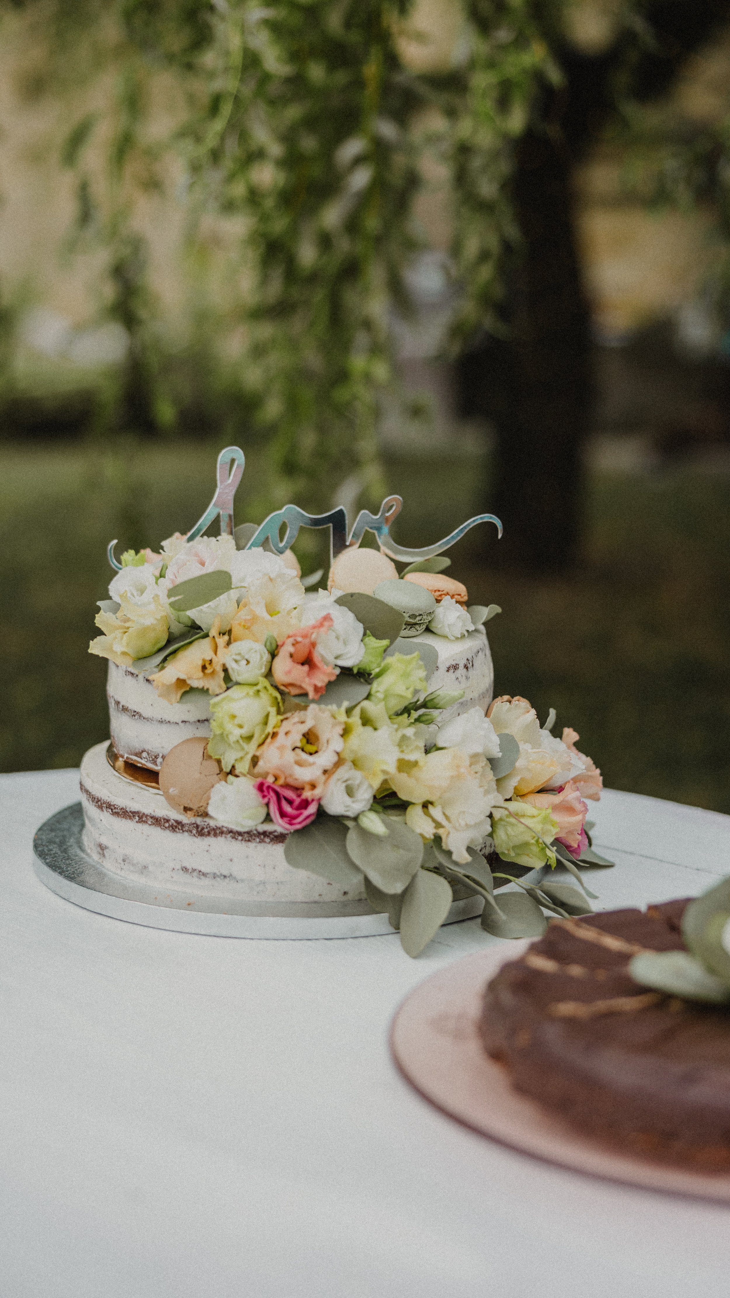 Malý „naked“ svatební dort zdobený pastelovými květy a nápisem „love“, položený na bílém stole venku.