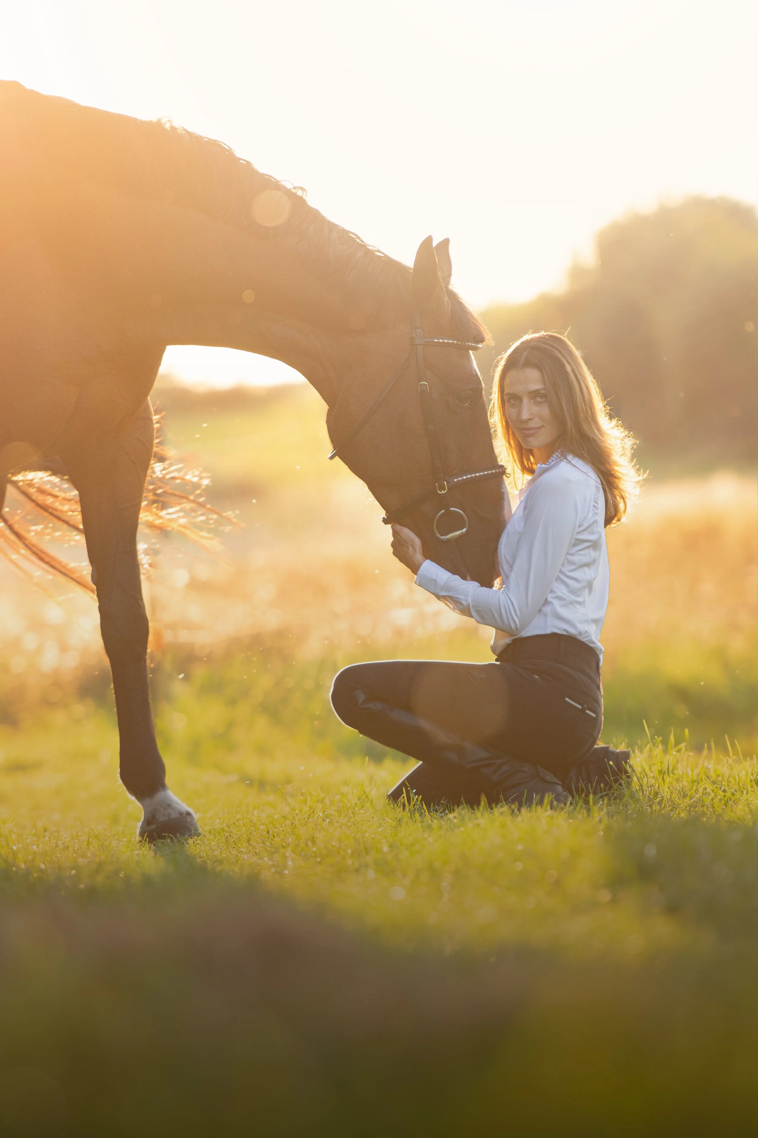 Žena klečí na trávě a jemně drží hlavu hnědého koně v prosluněném venkovním prostředí.