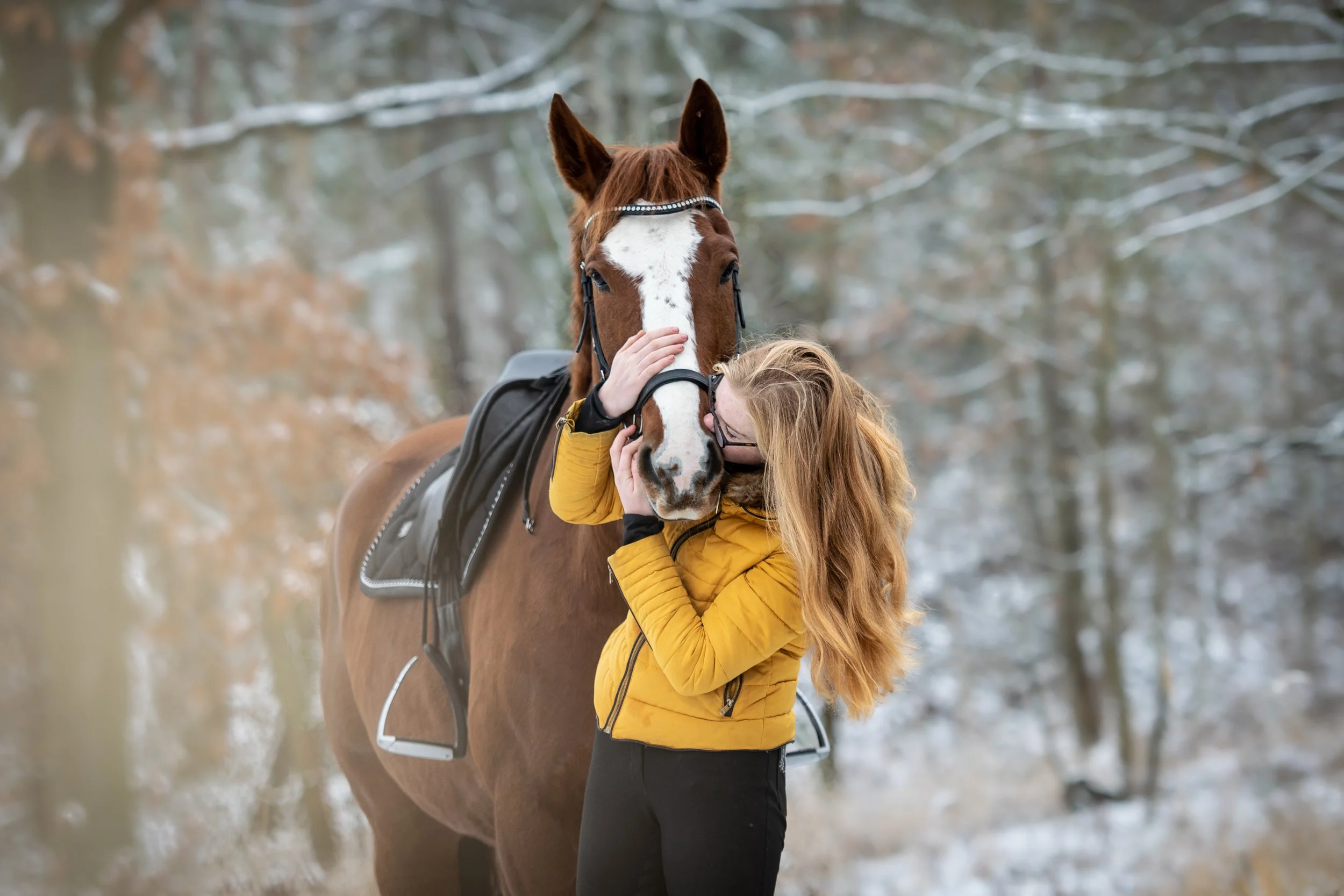Mladá žena s dlouhými zrzavými vlasy a brýlemi ve žluté bundě objímá a líbá hnědého koně s bílou lysinou, v zimním lese se sněhem na zemi a holými stromy.