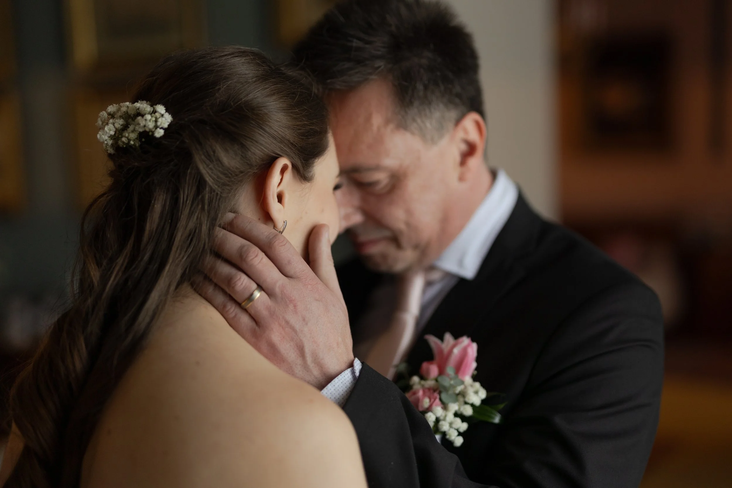 Pár sdílí intimní okamžik, dotýkají se čely a mají zavřené oči, oblečeni ve svatebním oblečení. Žena má ve vlasech květiny a prsten na ruce, muž má korsáž a snubní prsten.