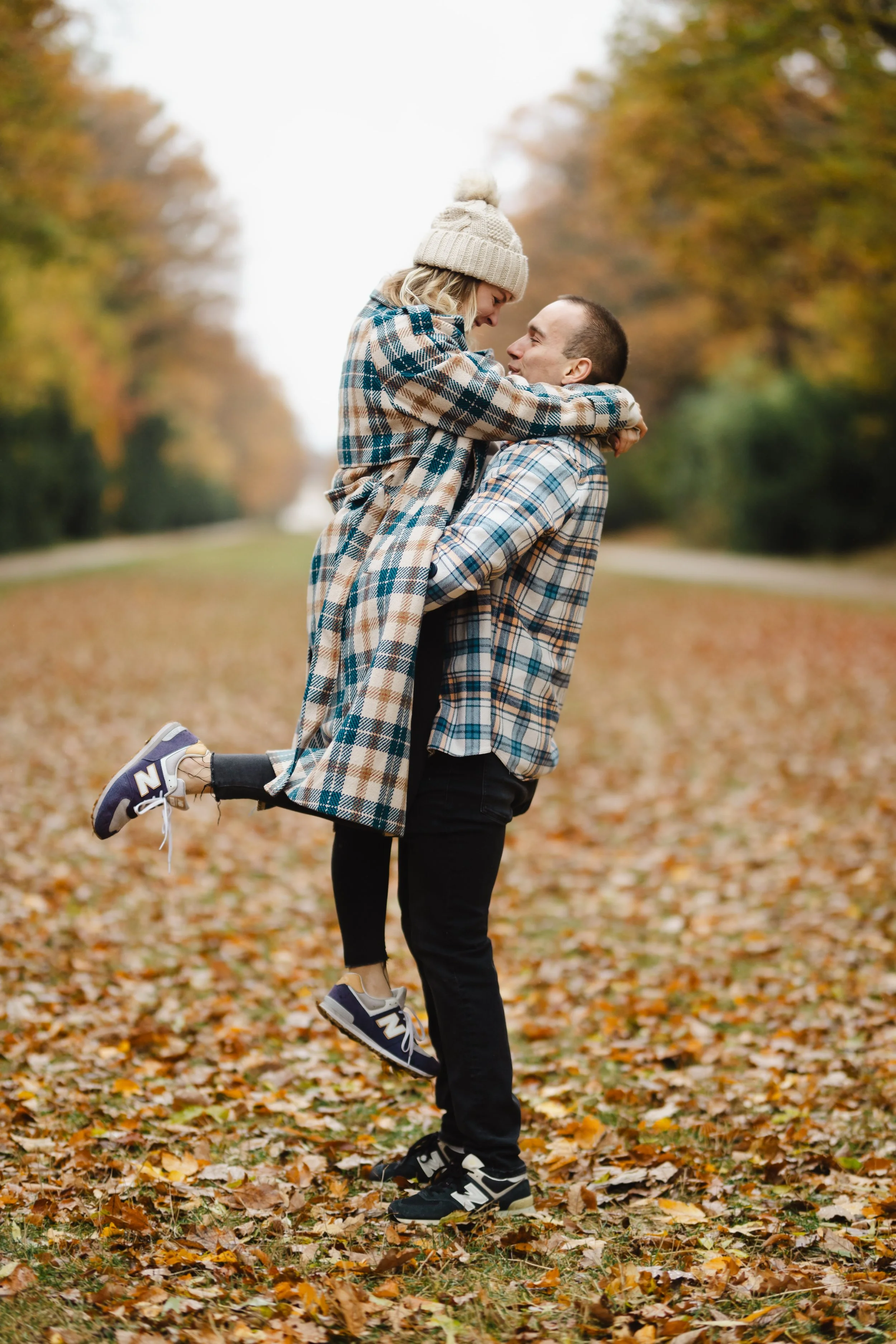Muž zvedající ženu v parku během podzimu mezi spadaným listím; oba mají na sobě kostkované košile.