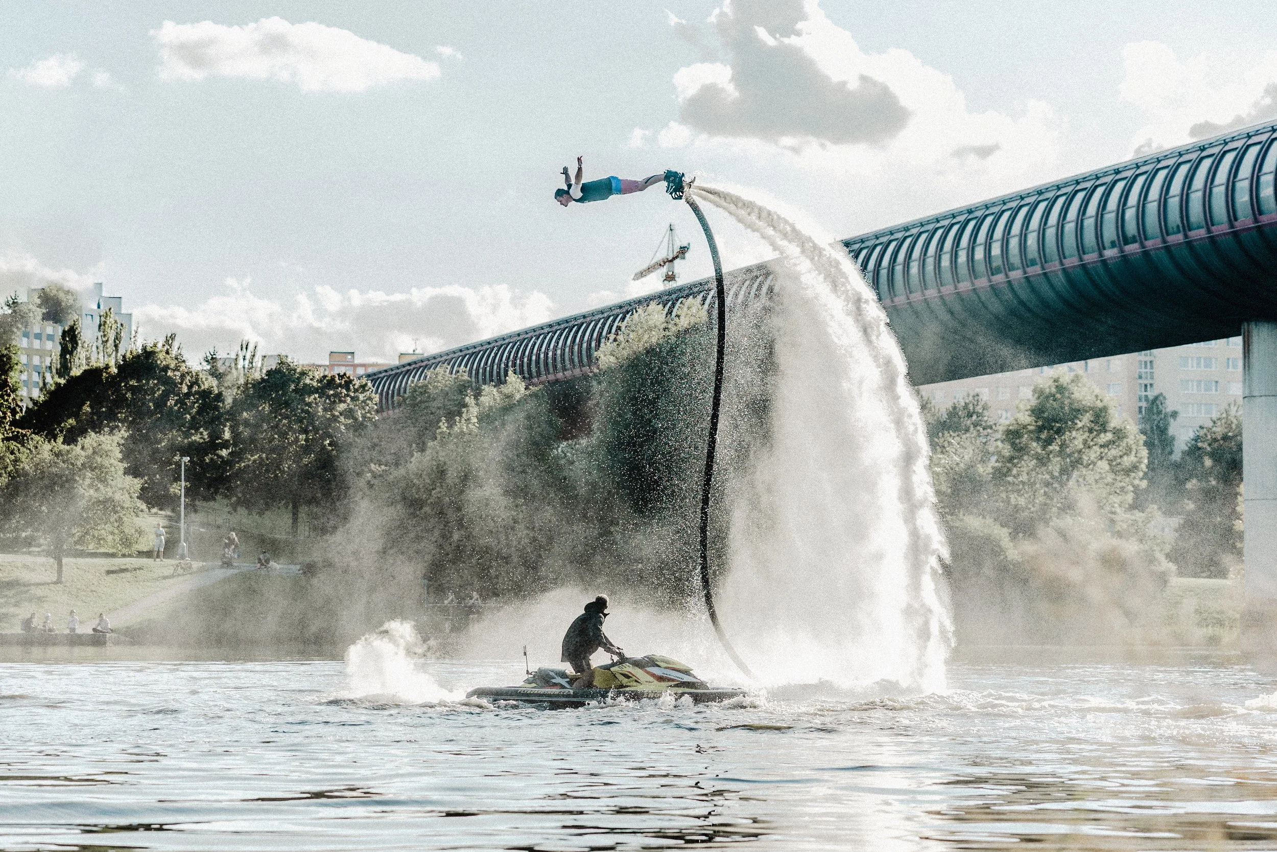 Osoba předvádí trik na flyboardu nad řekou, pod ní je obsluha na vodním skútru a v pozadí městská krajina se stromy a mostem.