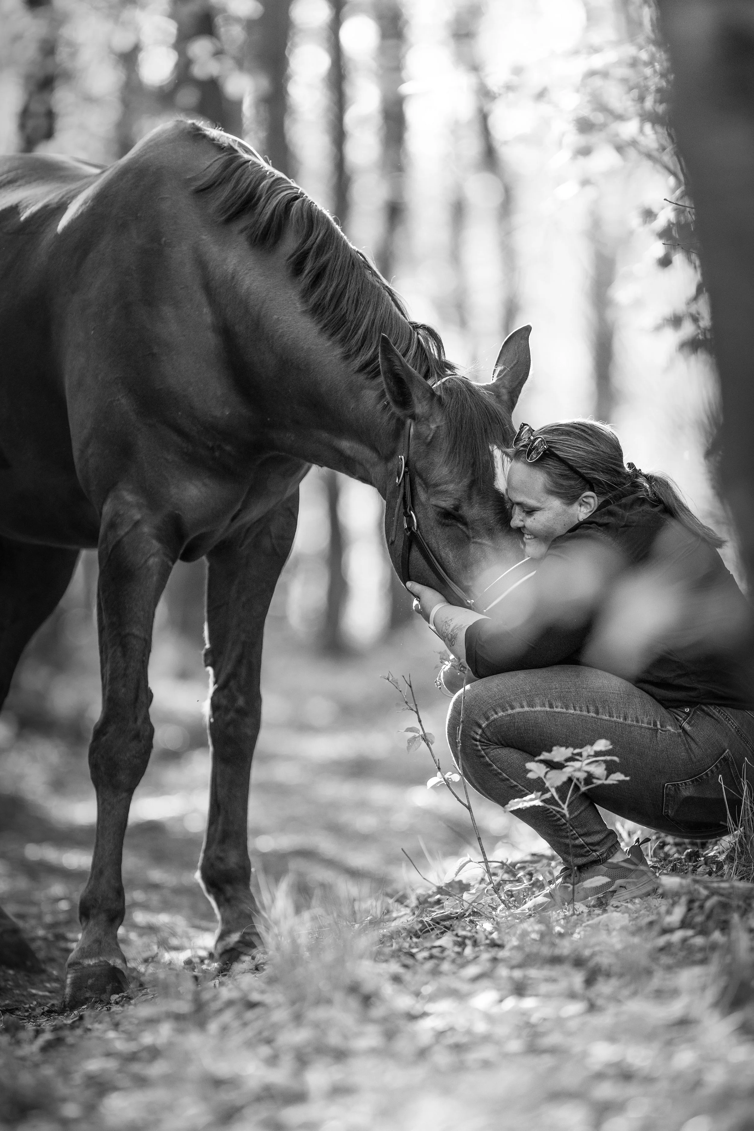 Žena objímající osedlaného koně v lese se stromy a slunečním světlem v pozadí, černobíle.