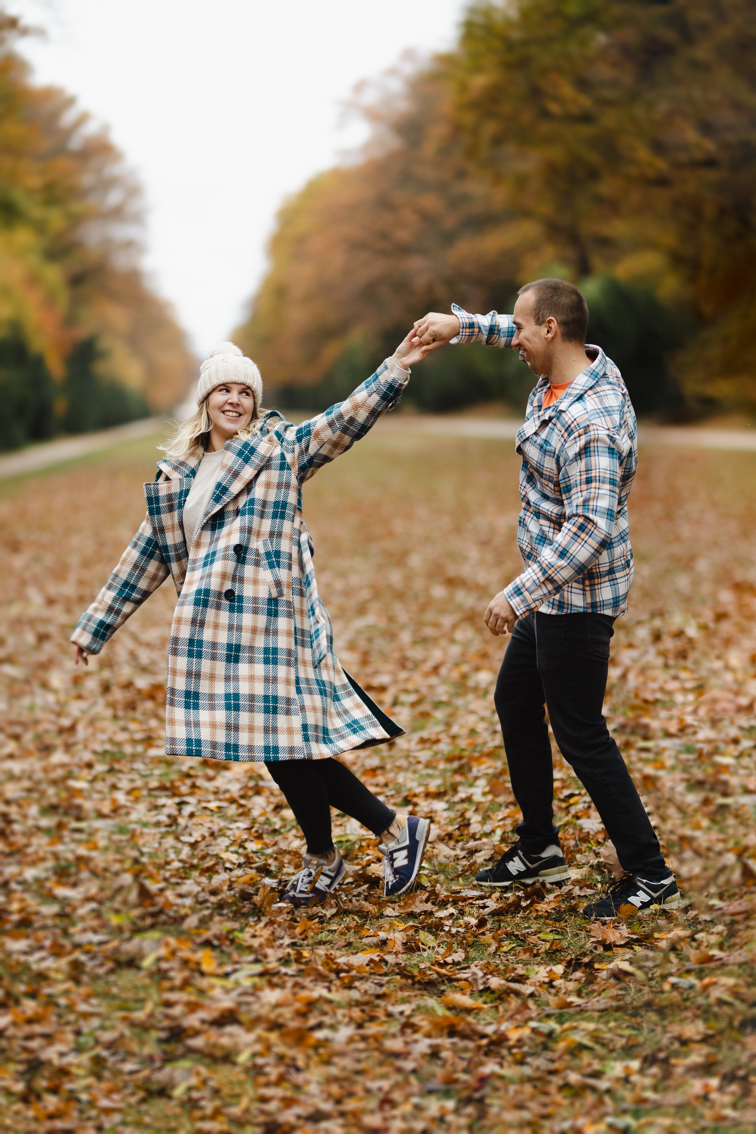 A smiling couple twirling in a park covered with fallen autumn leaves, wearing plaid jackets, with colorful fall trees in the background.