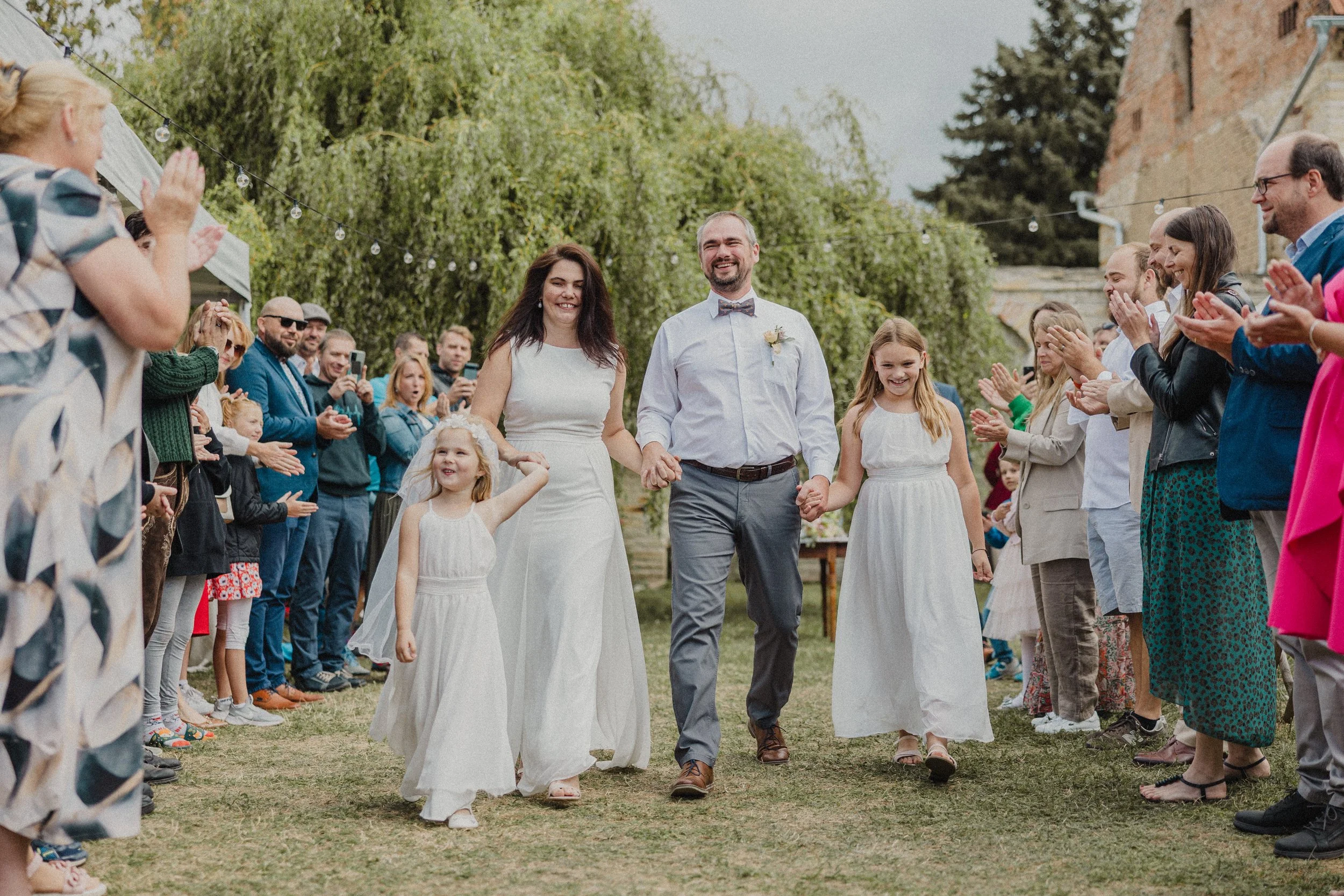 Nevěsta a ženich jdou ruku v ruce venkovní uličkou, obklopeni svatebními hosty, kteří tleskají a usmívají se.