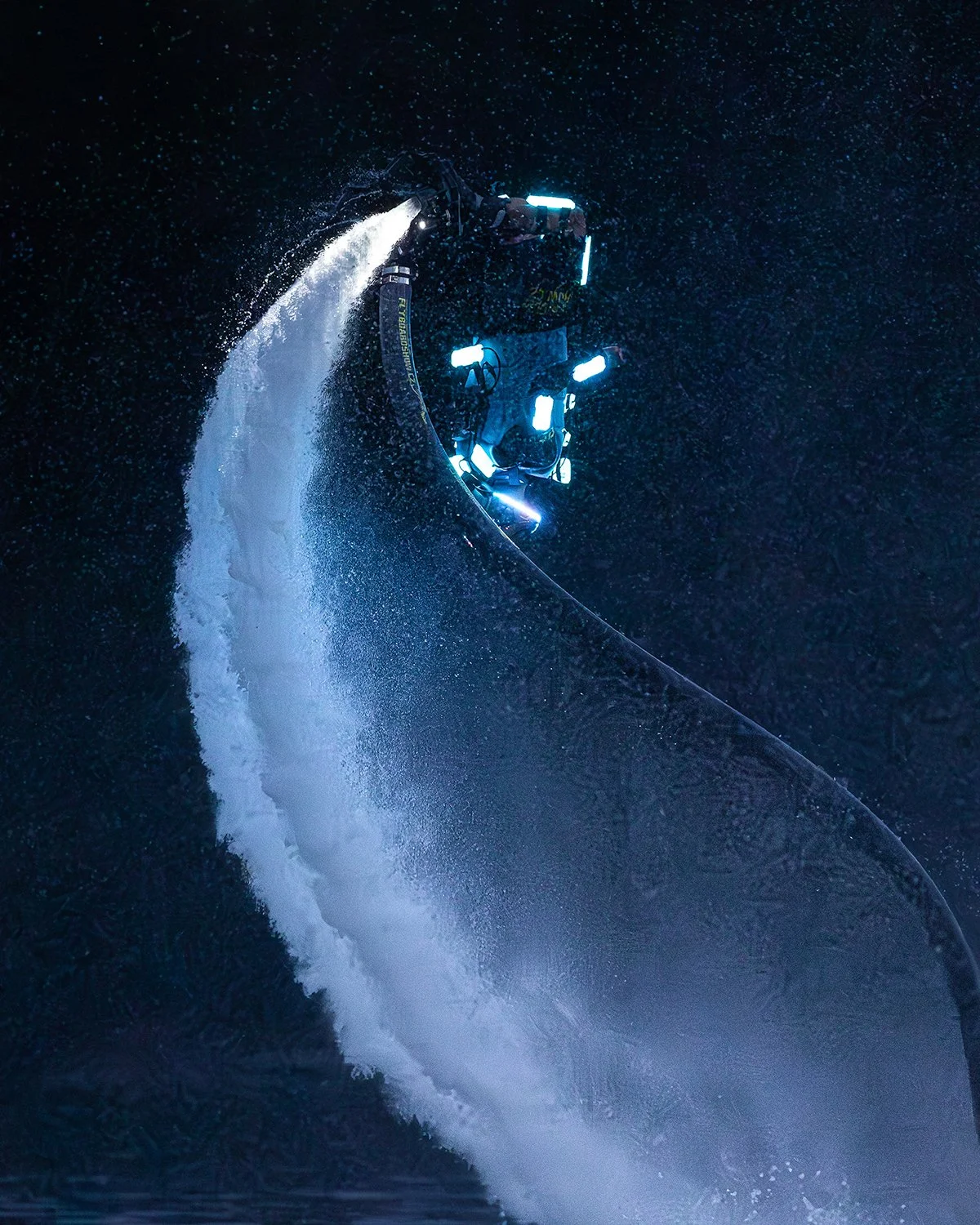 Osoba s čelovkou a výstrojí jede na vodním skútru ve vysoké rychlosti a za ní se zvedá velký vodní gejzír, v tmavém nočním prostředí.