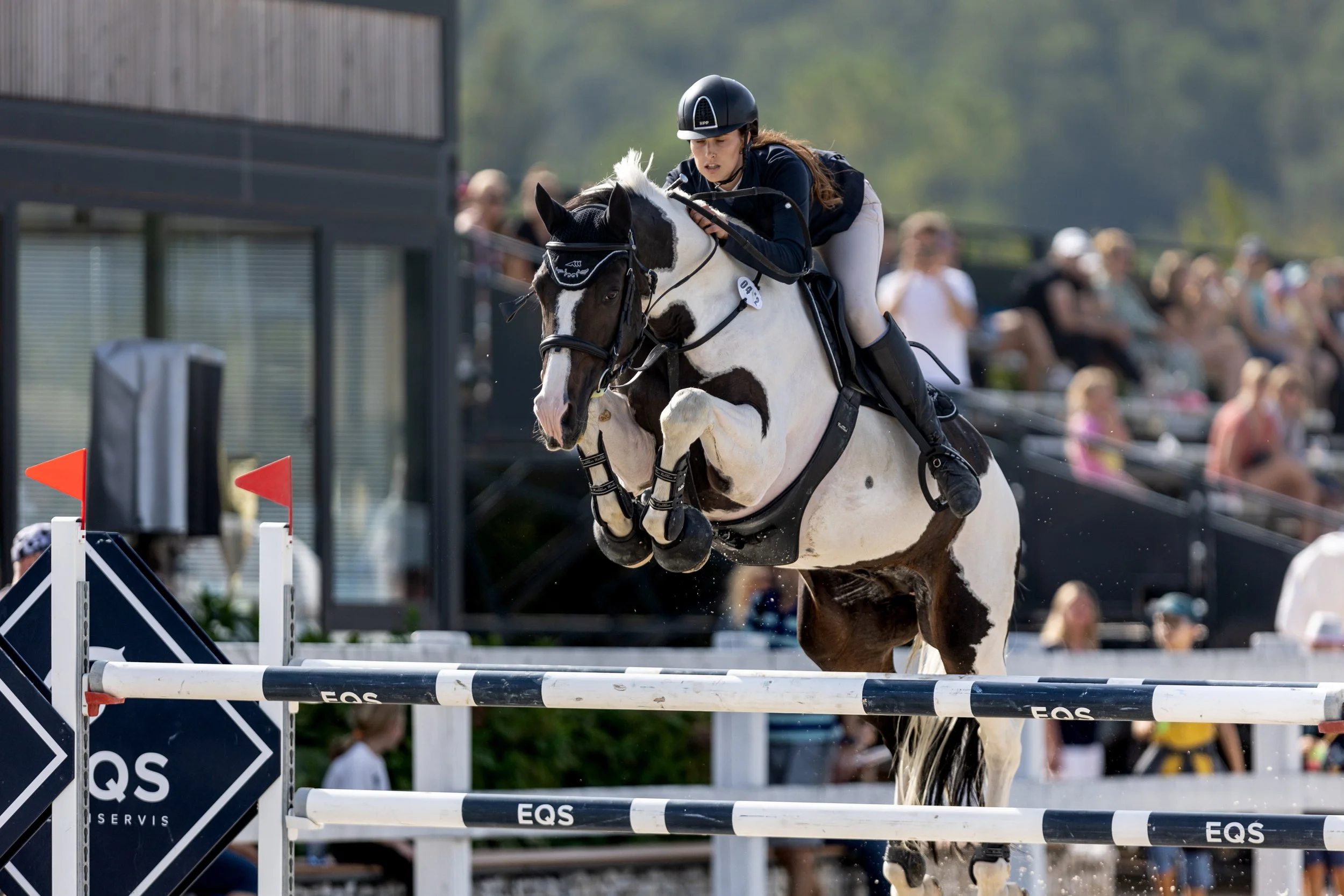 Jezdkyně na koni skáče přes překážku během parkurové soutěže, v pozadí přihlížejí diváci.