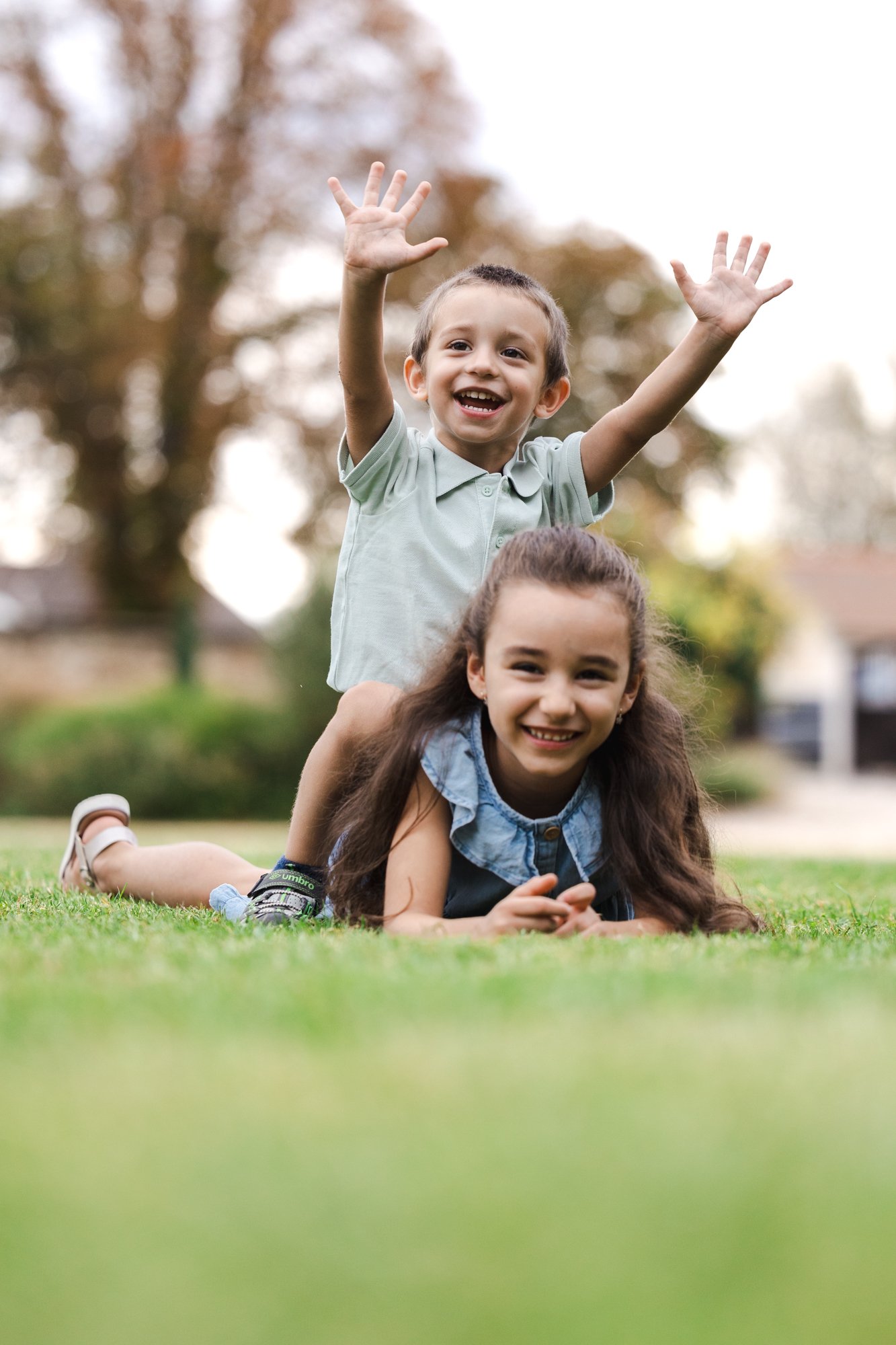 Chlapec a dívka si hrají venku na zelené trávě, v pozadí jsou stromy. Chlapec sedí dívce na zádech s rukama nahoře a usmívá se, zatímco dívka leží na břiše.