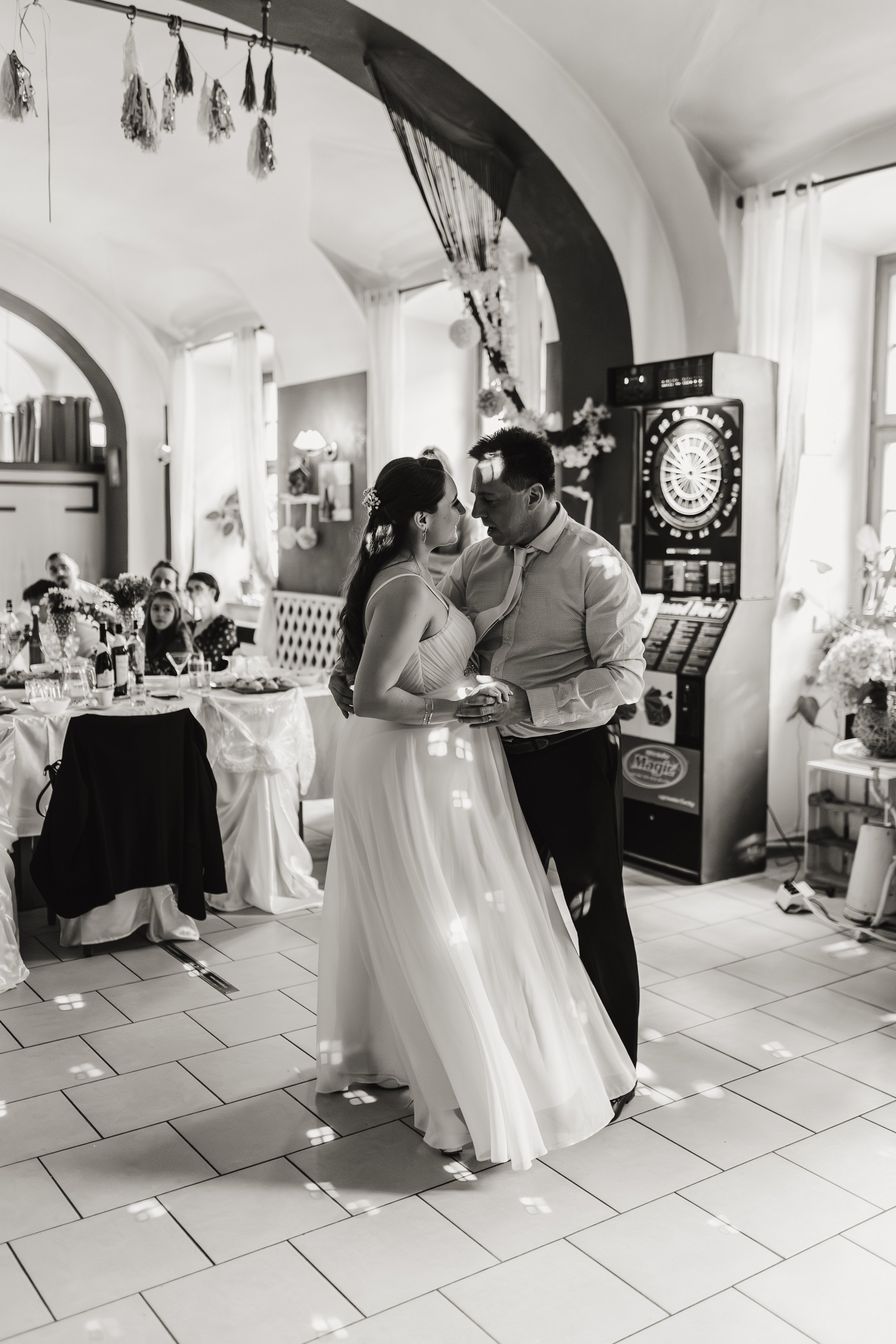 Nevěsta a ženich tančí na své svatební hostině ve vyzdobeném interiéru, zatímco hosté přihlížejí.