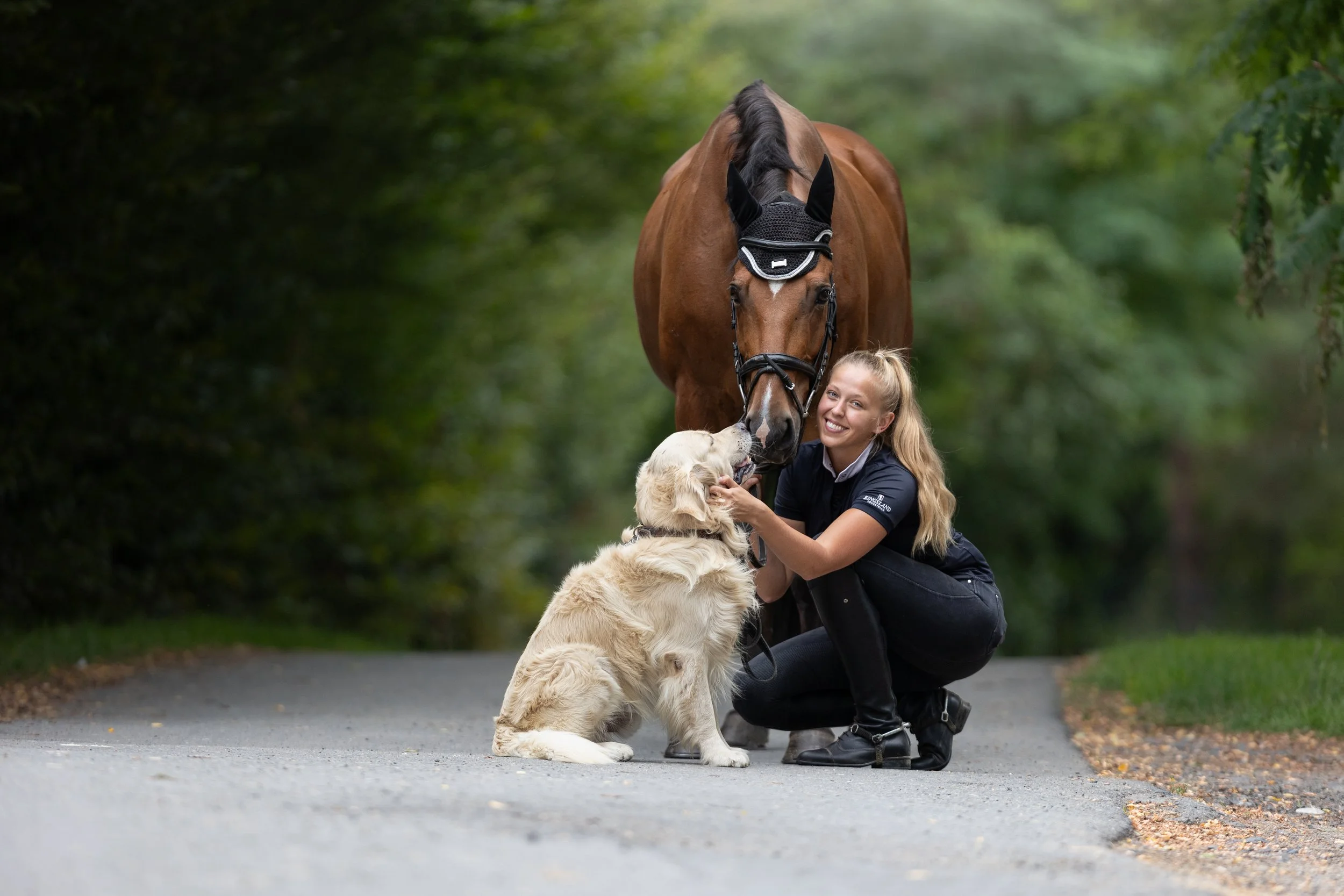 Mladá žena klečí na cestě, usmívá se a je se štěnětem zlatého retrívra a hnědým koněm. Kůň se tulí ke štěněti a žena drží štěně za hlavu. V pozadí jsou zelené stromy.
