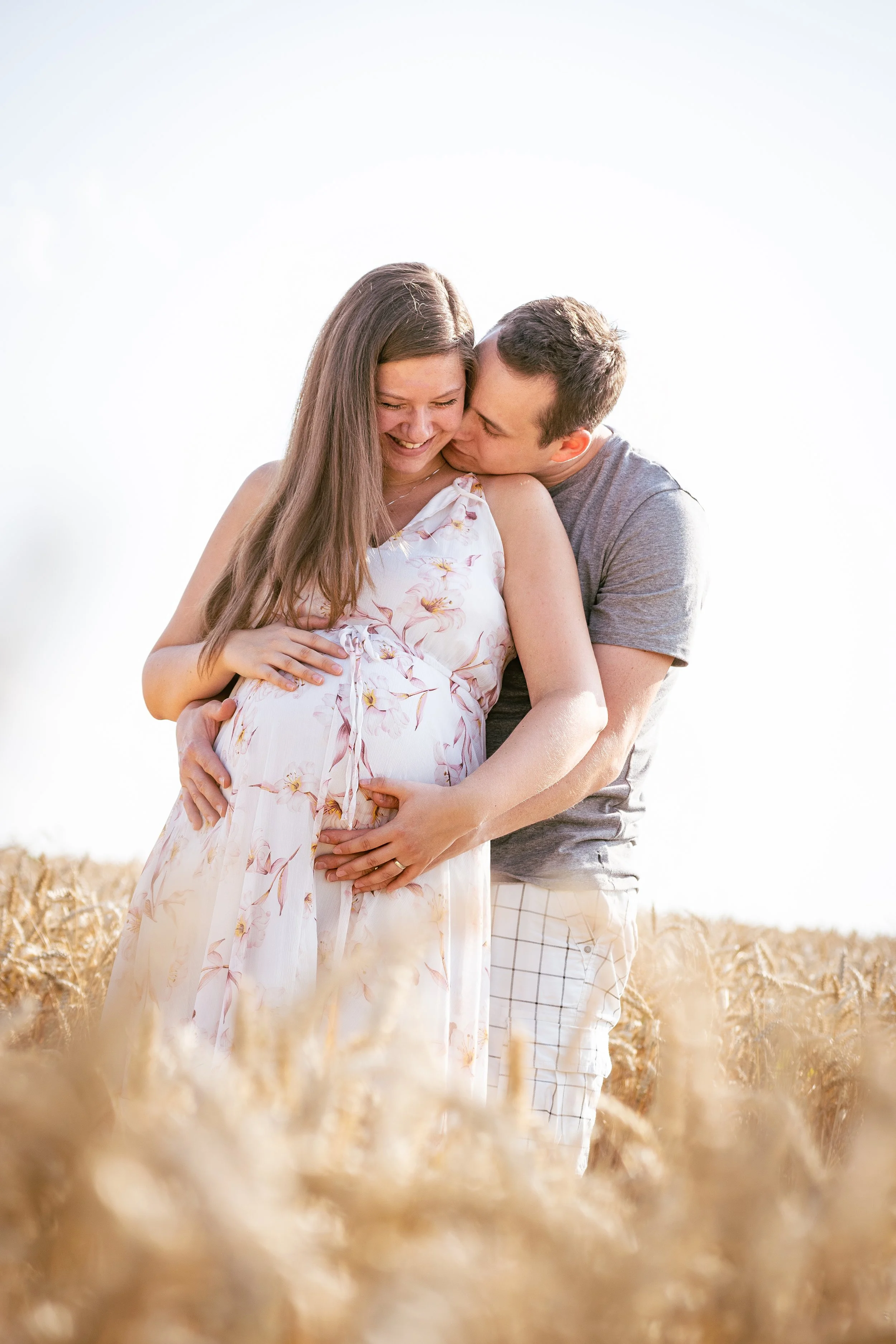 Těhotná žena a její partner stojí v pšeničném poli, usmívají se a dotýkají se jejího bříška, zatímco muž ji líbá na tvář.