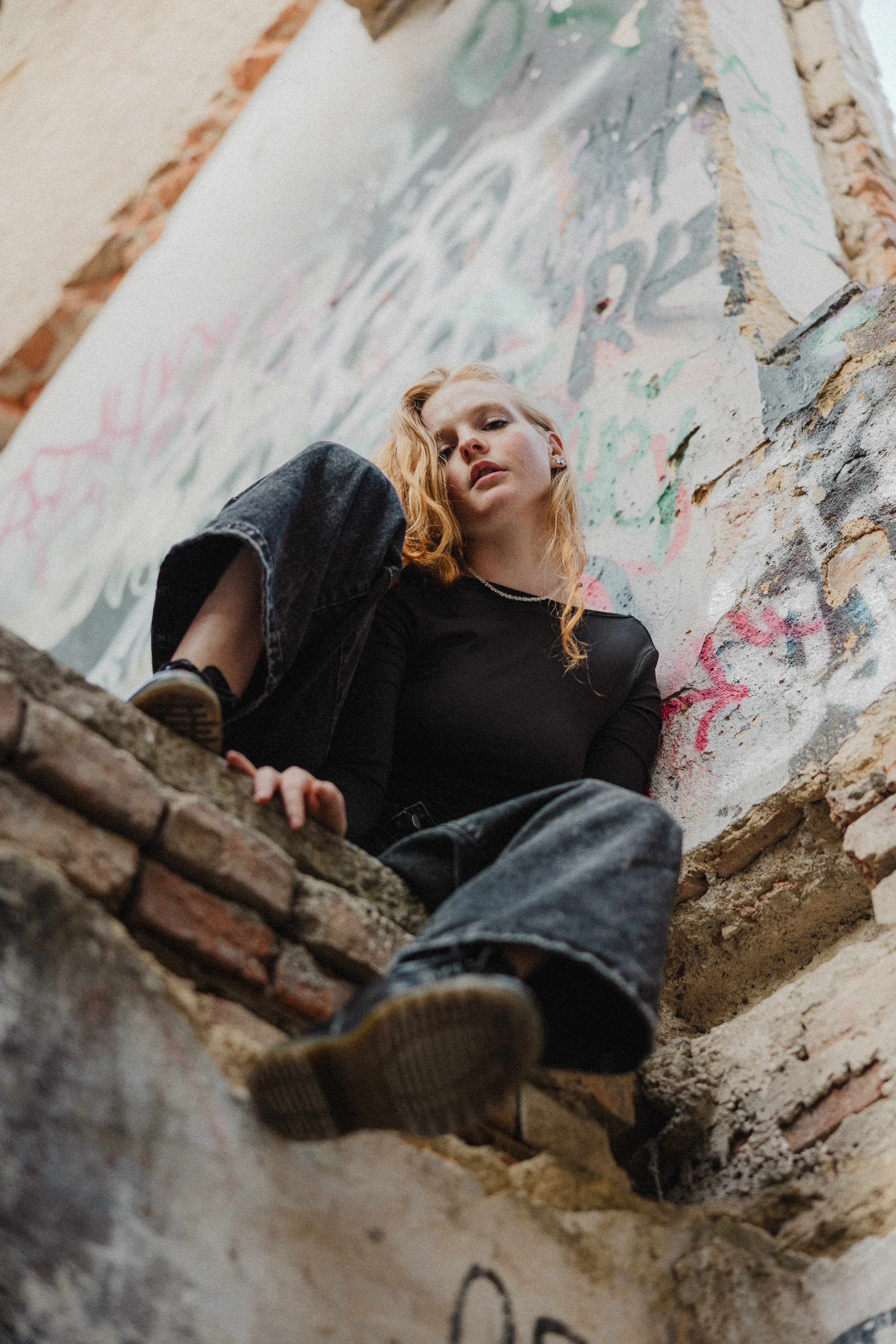 Mladá žena s načervenalými kudrnatými vlasy v černém topu s dlouhým rukávem a tmavých džínách, sedící na cihlové zídce.