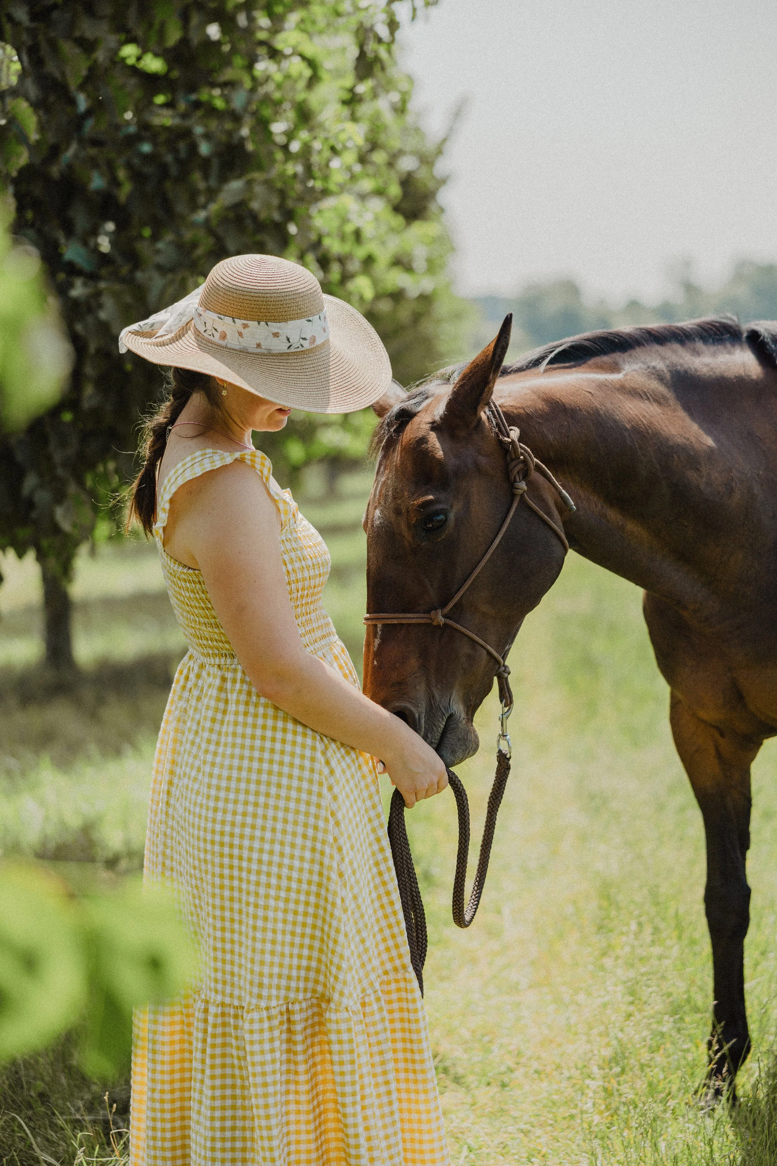 Žena ve žlutých kostkovaných šatech a klobouku se širokou krempou stojí vedle hnědého koně na travnaté louce se stromy v pozadí.