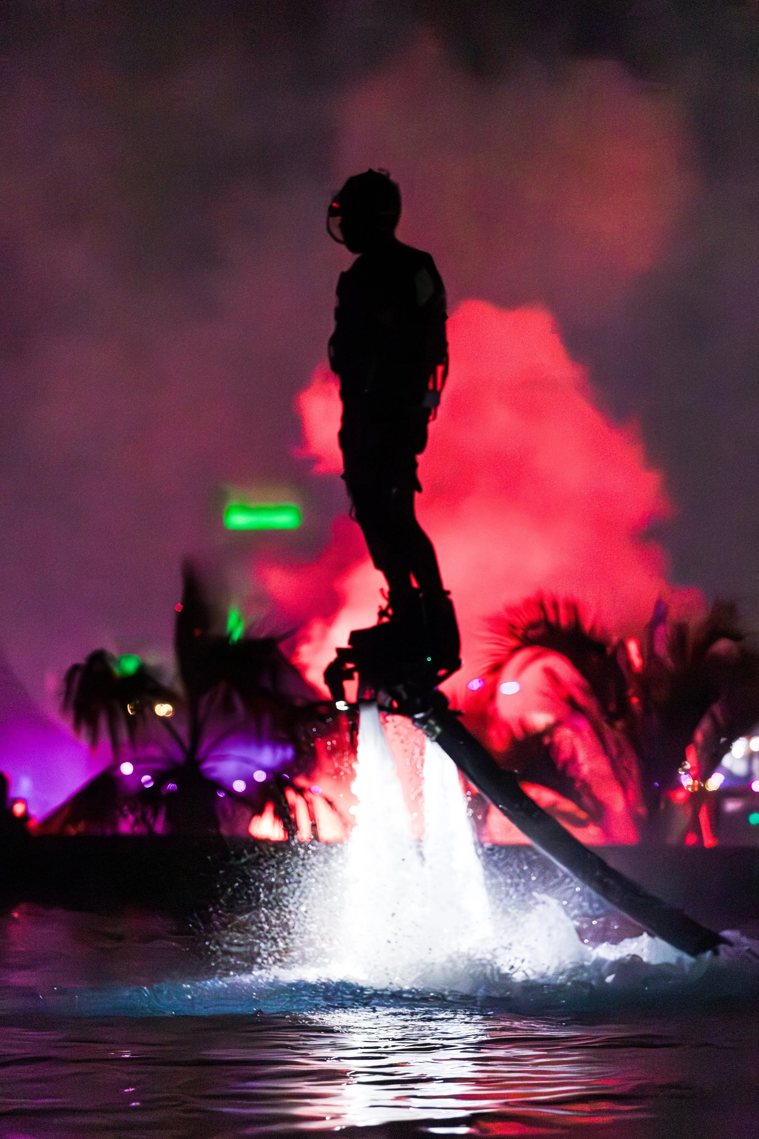 Osoba jede na flyboardu v noci, s barevnými světly a kouřem v pozadí.