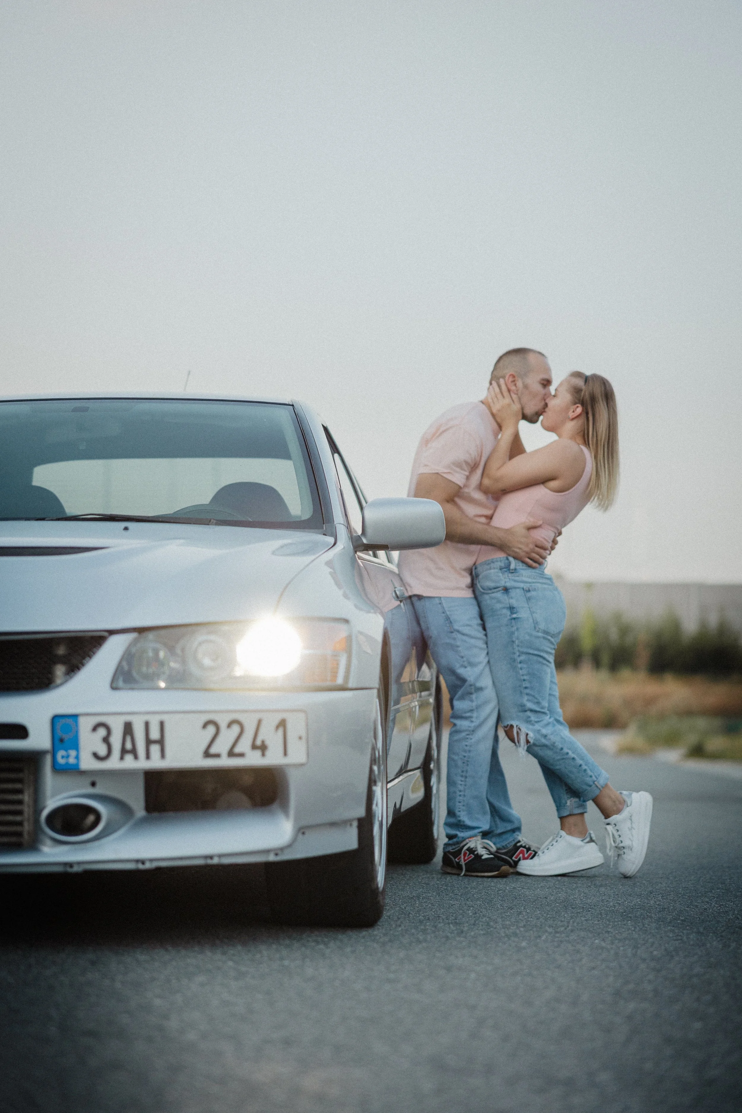 Pár, který se líbá a objímá vedle stříbrného auta na otevřené silnici, s jasnou oblohou v pozadí.