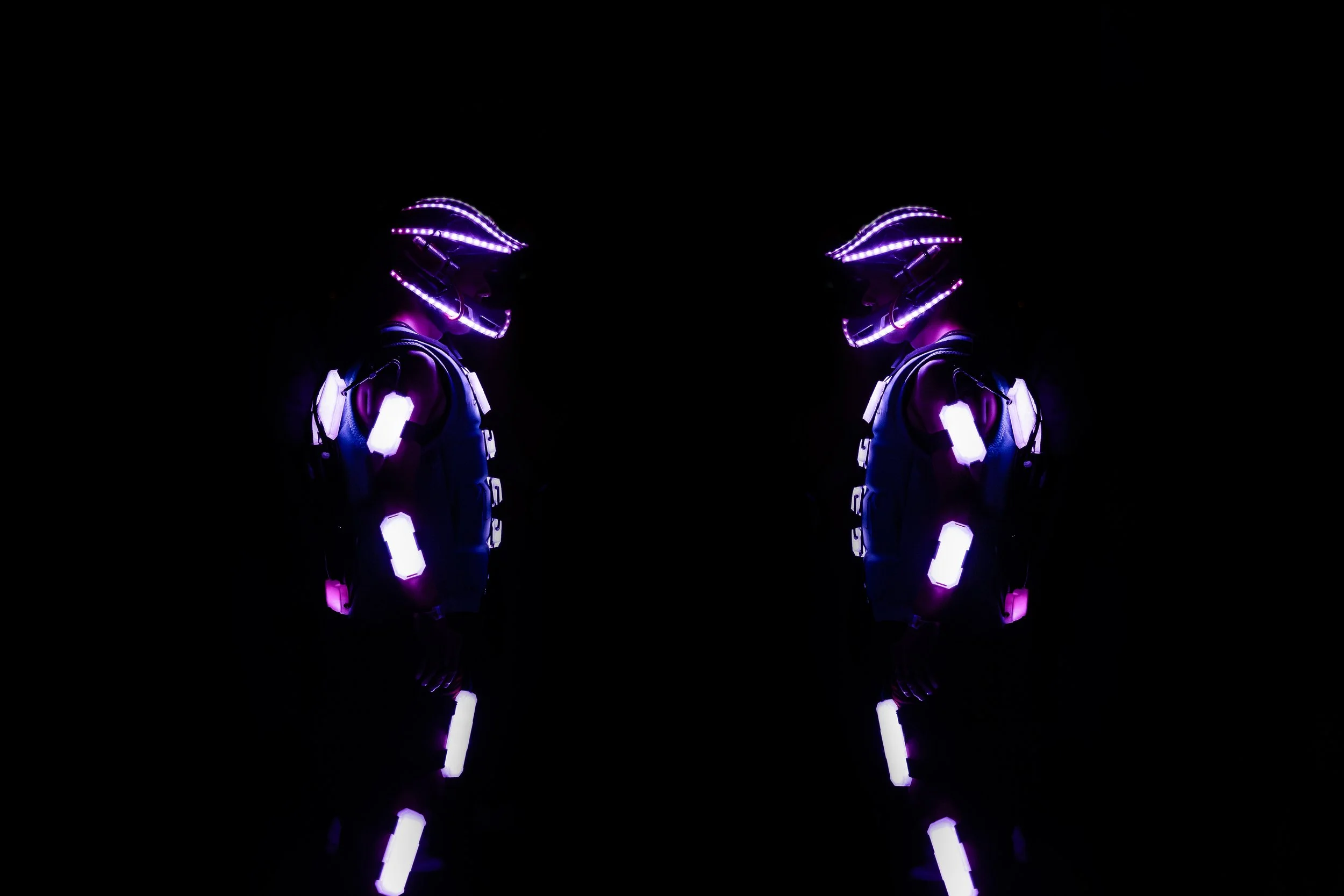 Dva lidé v futuristických, svítících LED oblecích a helmách stojí proti sobě v tmavém prostředí.