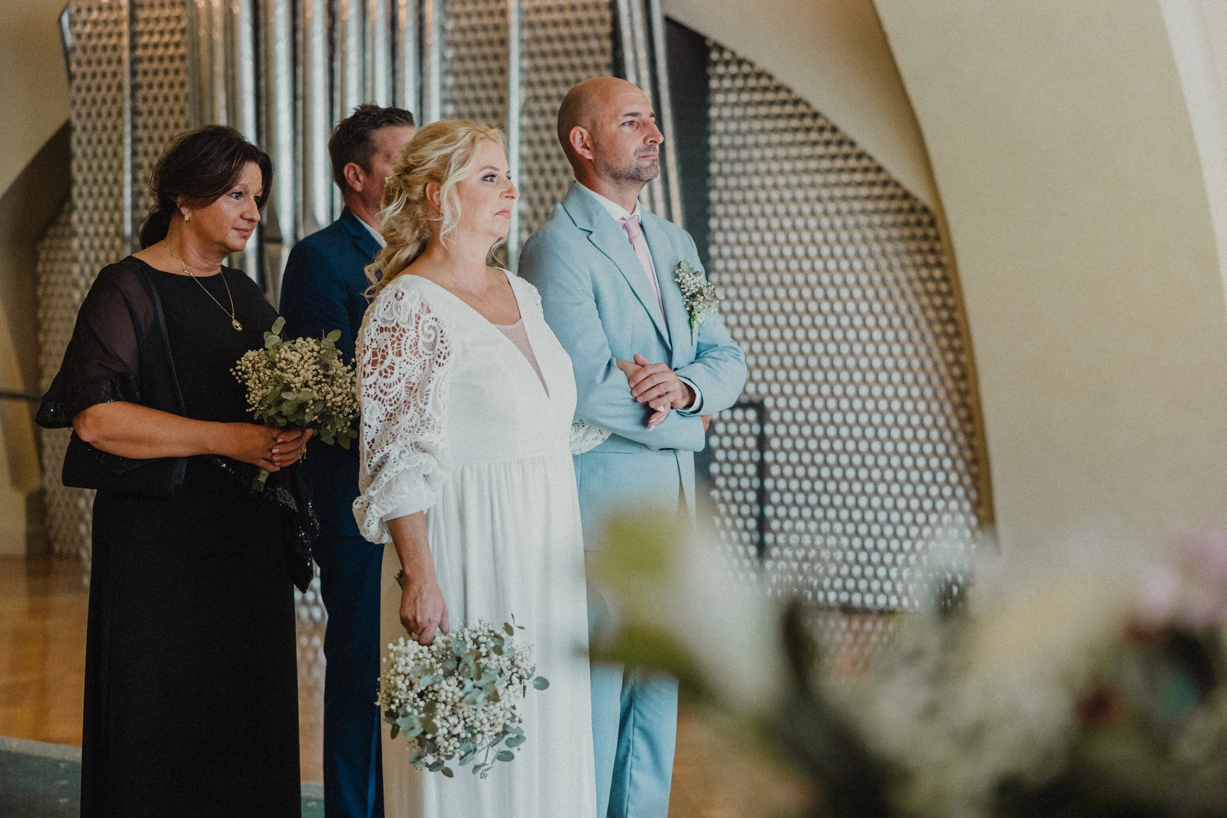 Skupina pěti lidí stojí v řadě během svatebního obřadu; nevěsta drží kytici a má bílé šaty s krajkovými detaily, ženich má světle modrý oblek s korsáží.