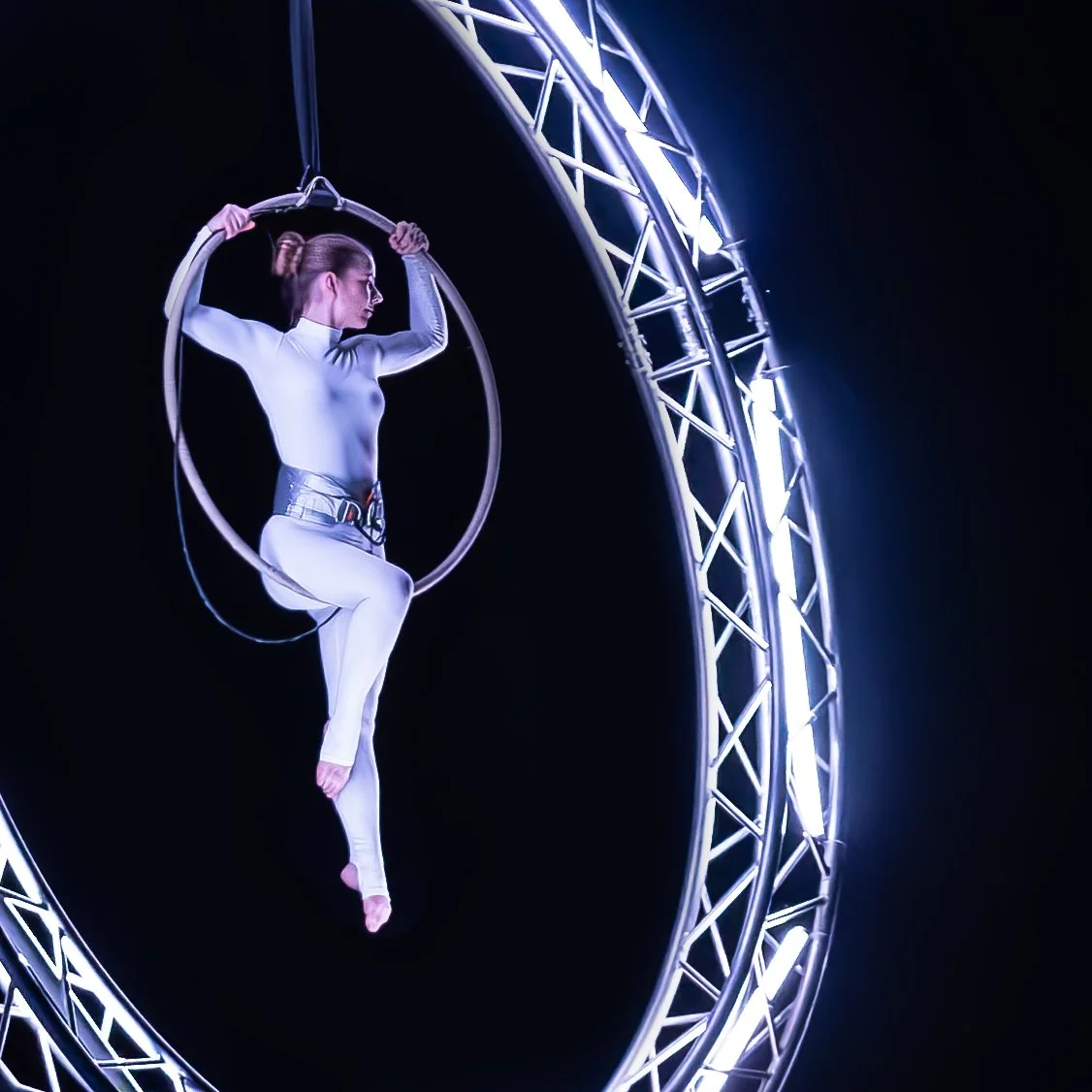 Akrobatka v bílém kostýmu předvádí vzdušnou akrobacii na kruhu uvnitř kruhové kovové konstrukce s jasnými bílými světly.