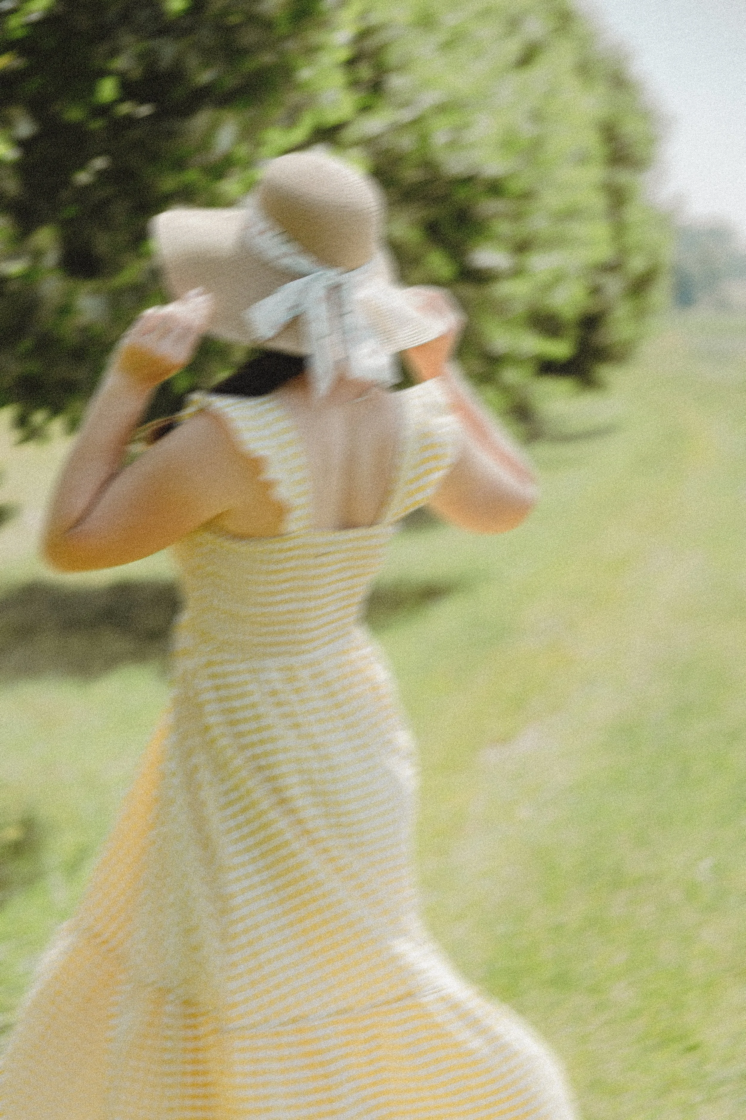 Žena ve žlutých pruhovaných šatech a klobouku se širokou krempou stojící venku poblíž zelených stromů nebo keřů, upravující si klobouk.