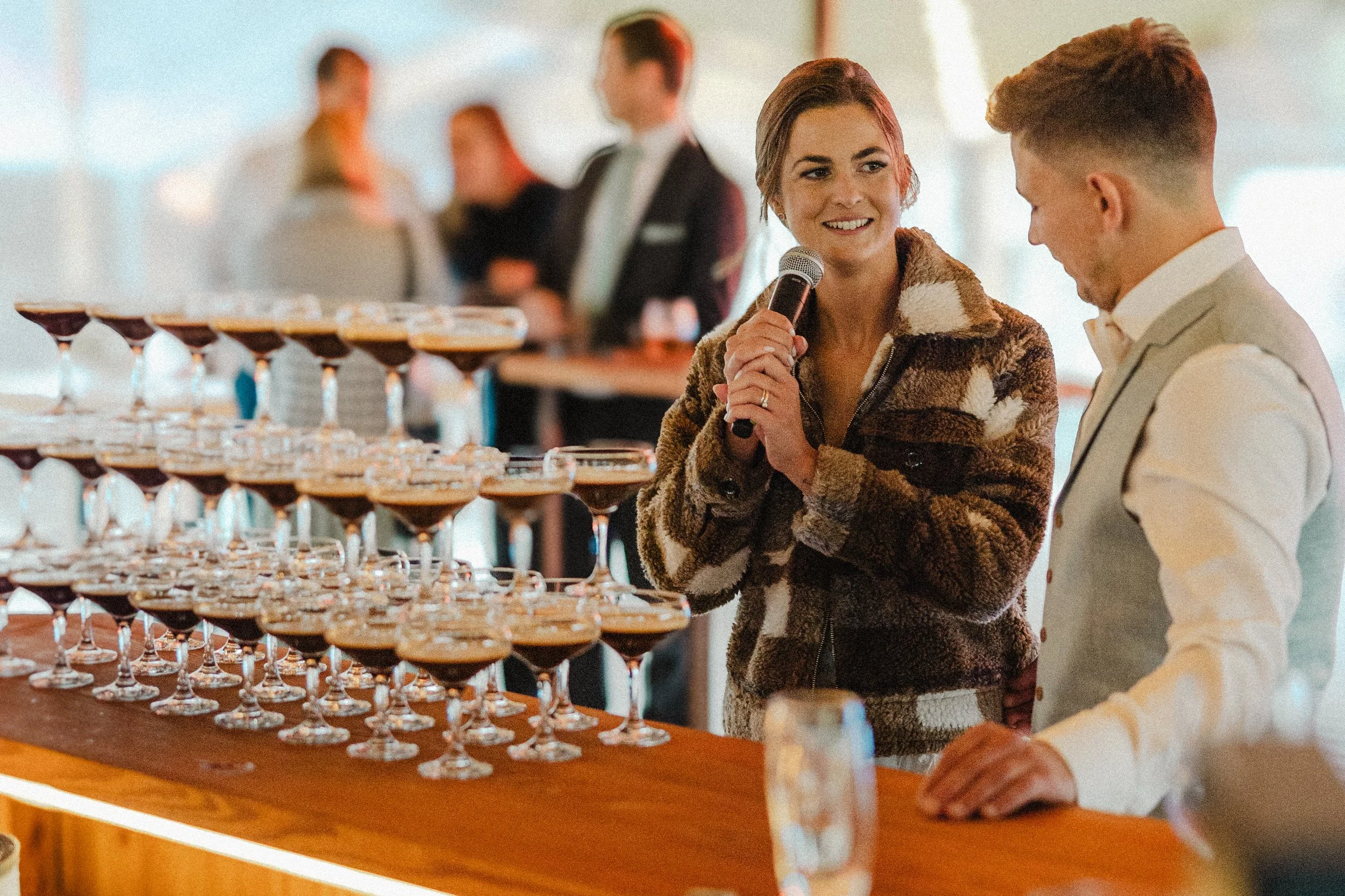 Žena s mikrofonem mluví s mužem u baru, na jehož pultu je řada koktejlových sklenic naplněných martini espresso. V pozadí jsou rozmazaní lidé na svatbě.