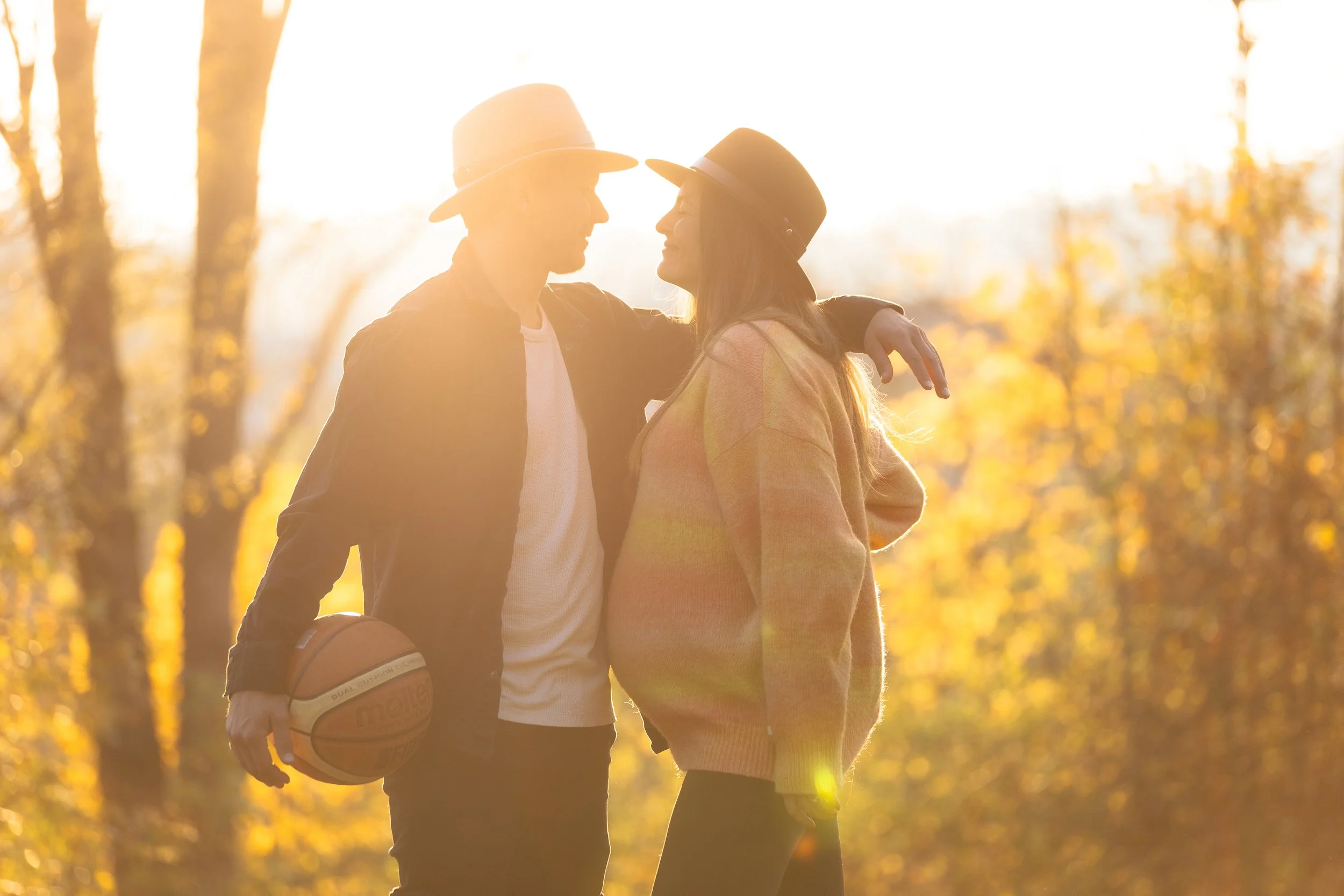 Muž a žena stojí těsně u sebe venku při západu slunce. Muž má ruku kolem ženy a v druhé drží basketbalový míč. Oba mají na sobě klobouky a ležérní oblečení, v pozadí jsou podzimní stromy.