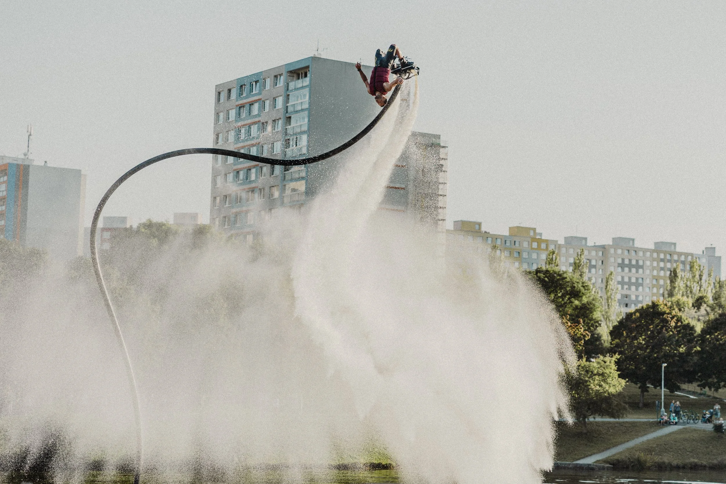Osoba na vodním jetpacku letí nad vodní hladinou, v pozadí jsou městské budovy.