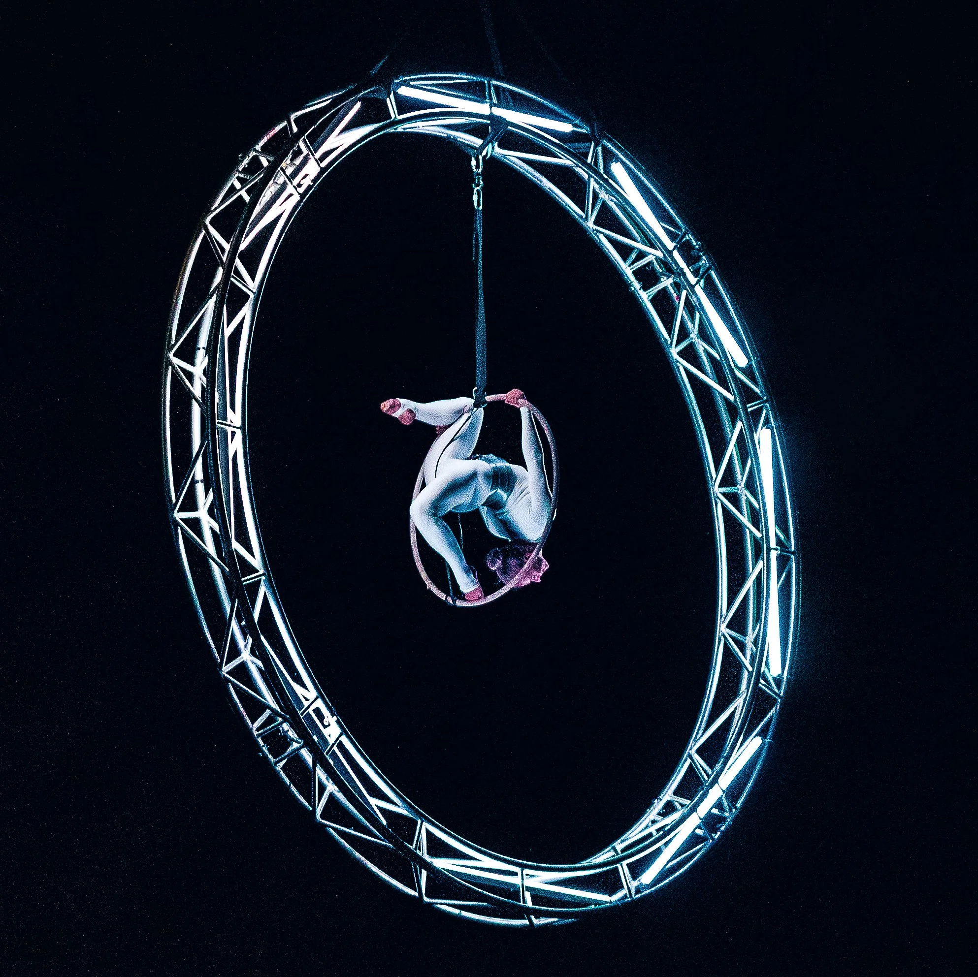 Akrobat visí hlavou dolů v akrobatické pozici uvnitř svítícího kruhového kovového kruhu na černém pozadí.