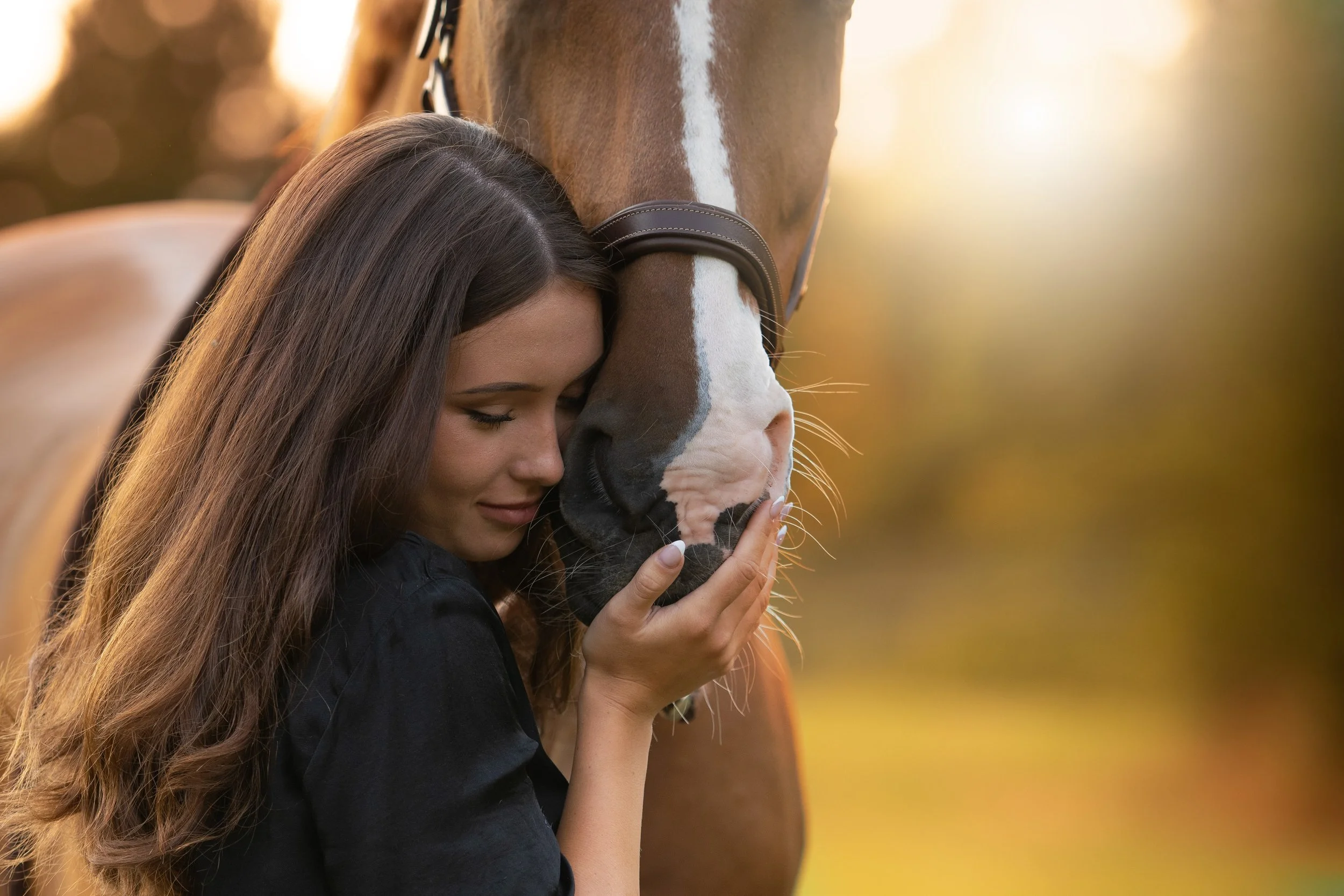 Mladá žena s dlouhými hnědými vlasy jemně drží koňskou hlavu a opírá svou hlavu o koňskou tvář, venku s teplým, rozostřeným pozadím.