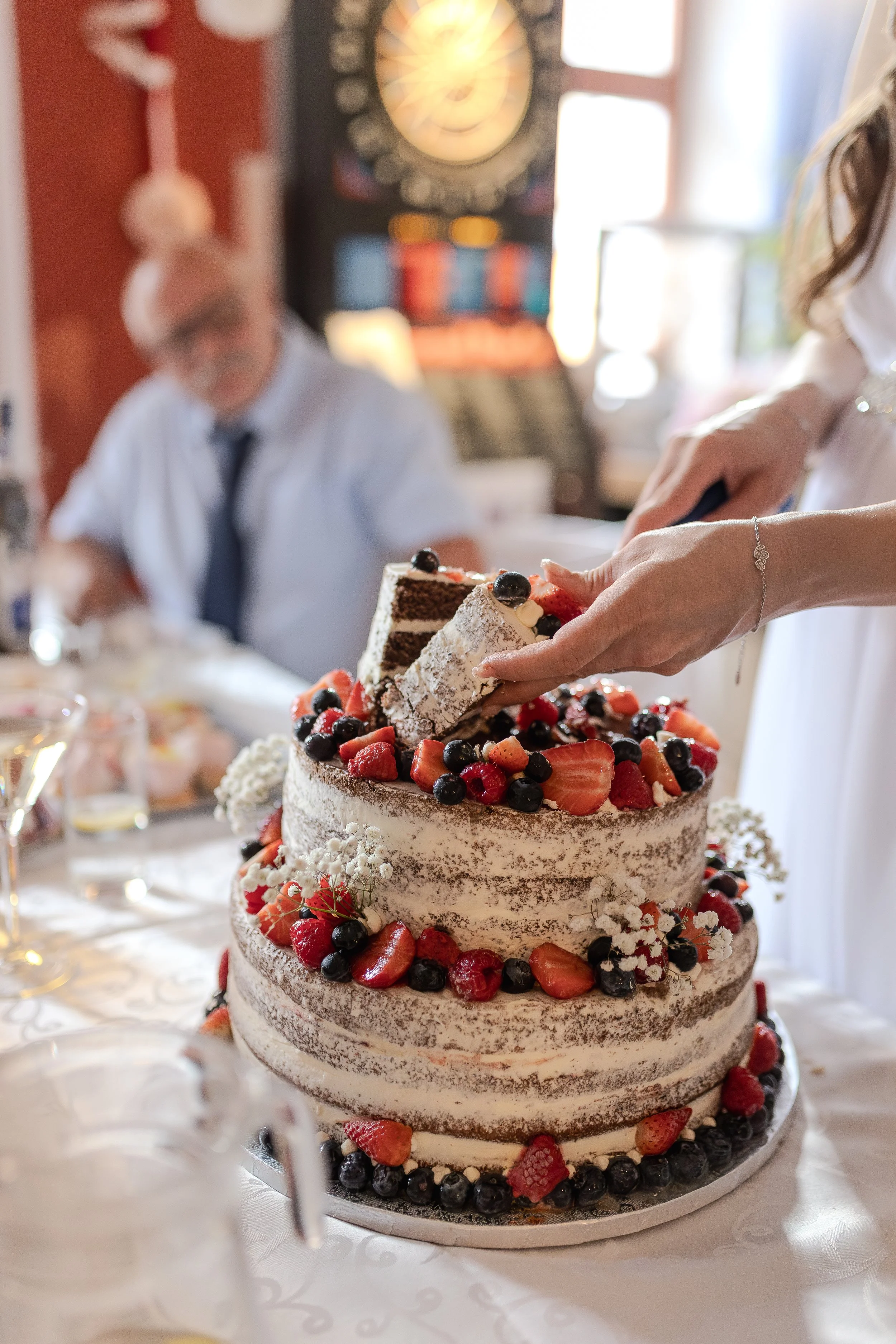 Osoba krájí třípatrový svatební dort zdobený jahodami, borůvkami a bílými květy; v pozadí sedí u stolu muž.