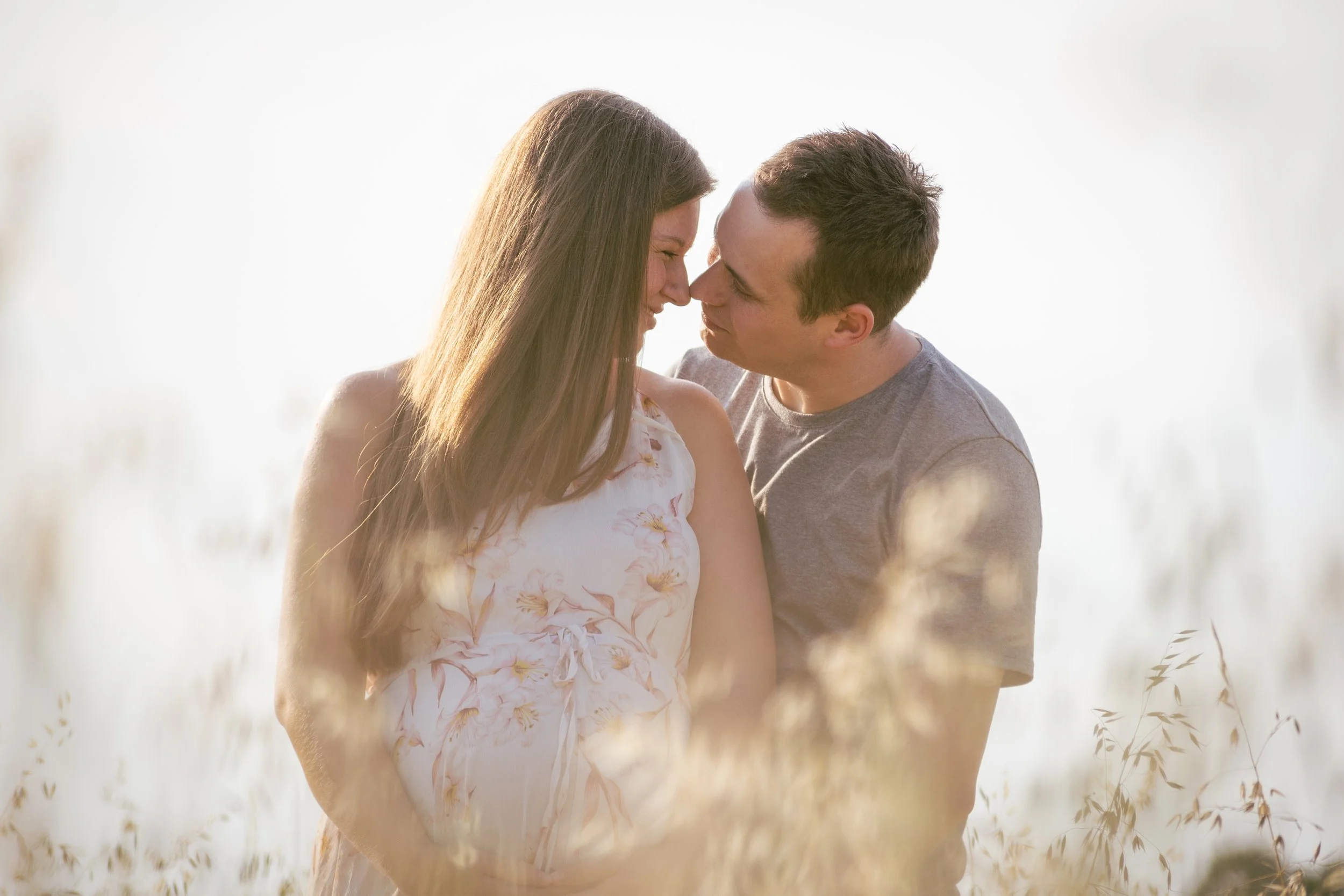 A couple standing close together in a field of tall grass, with their foreheads and noses touching, during a sunny day.