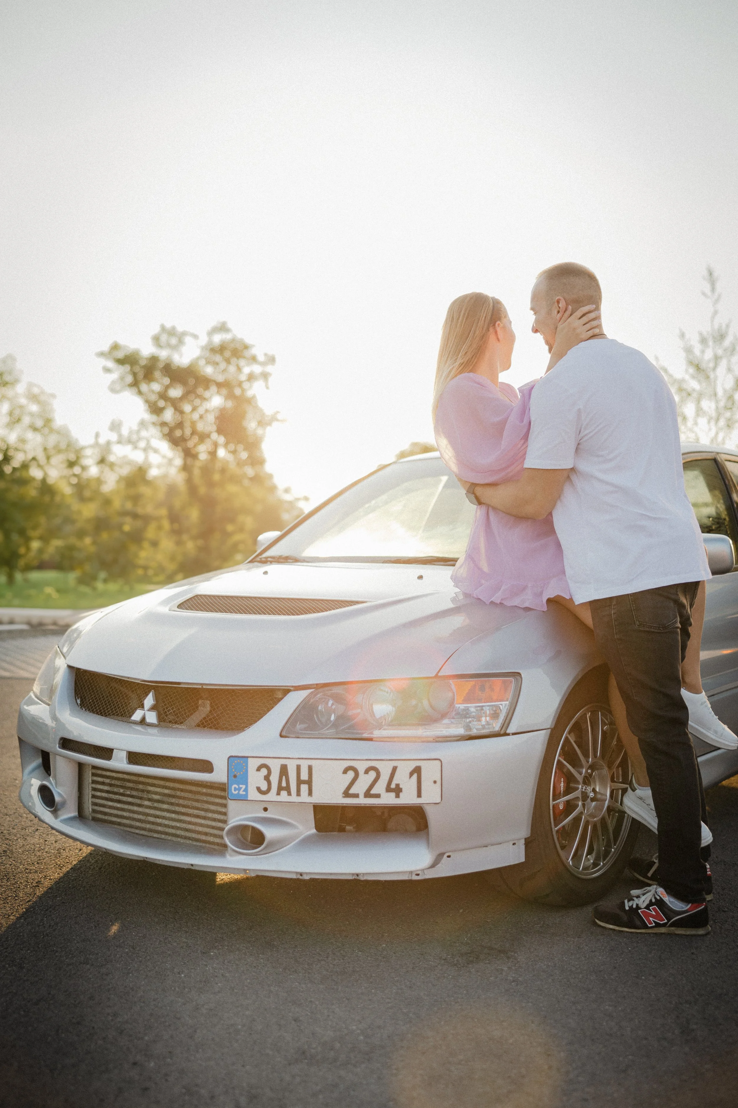 Pár stojící u stříbrného vozu Mitsubishi Lancer zaparkovaného u krajnice, objímající se a usmívající se na sebe při západu slunce, se stromy v pozadí.
