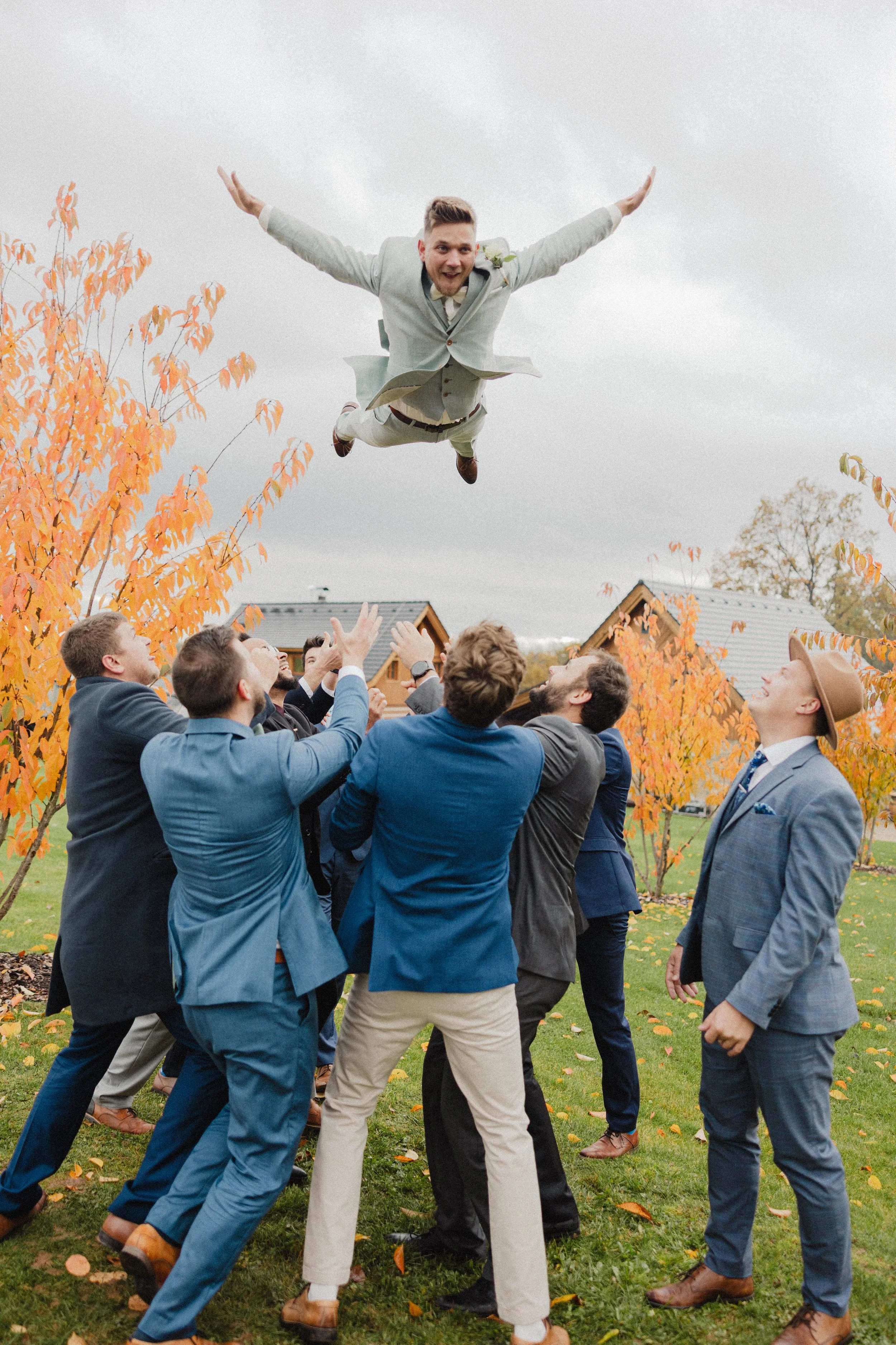 Muž ve světle šedém obleku je venku na podzim vyhazován do vzduchu skupinou mužů v oblecích.
