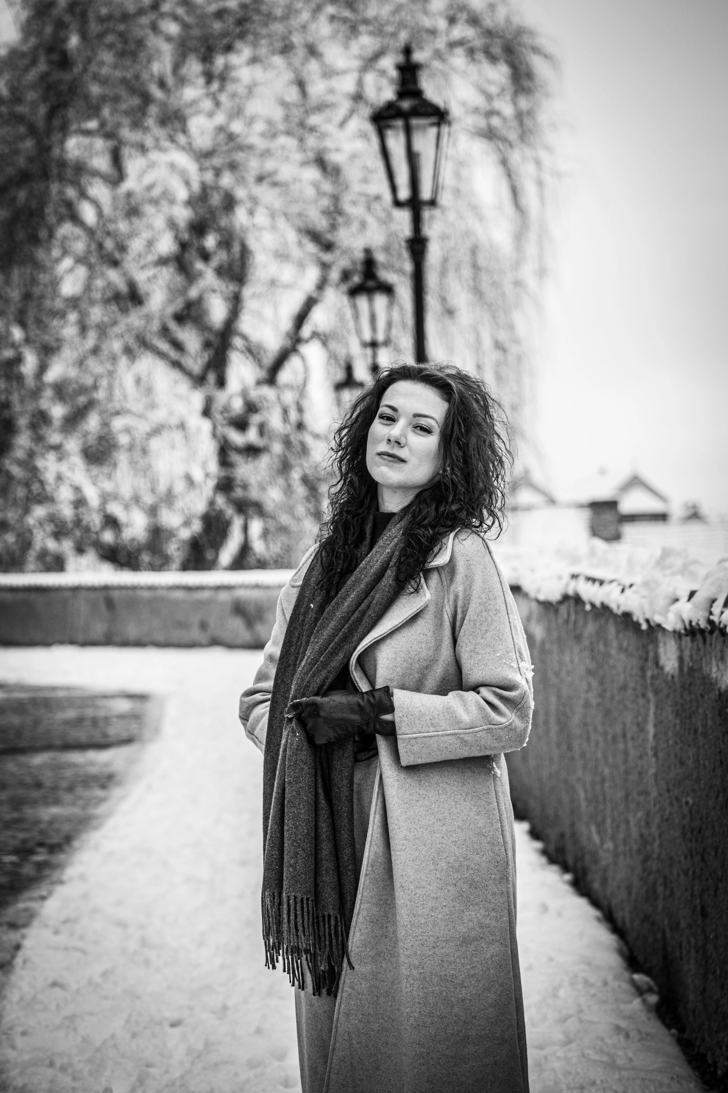 Žena s kudrnatými vlasy stojící venku na zasněžené cestě, oblečená v kabátu, rukavicích a šále; v pozadí jsou stromy a lampy.
