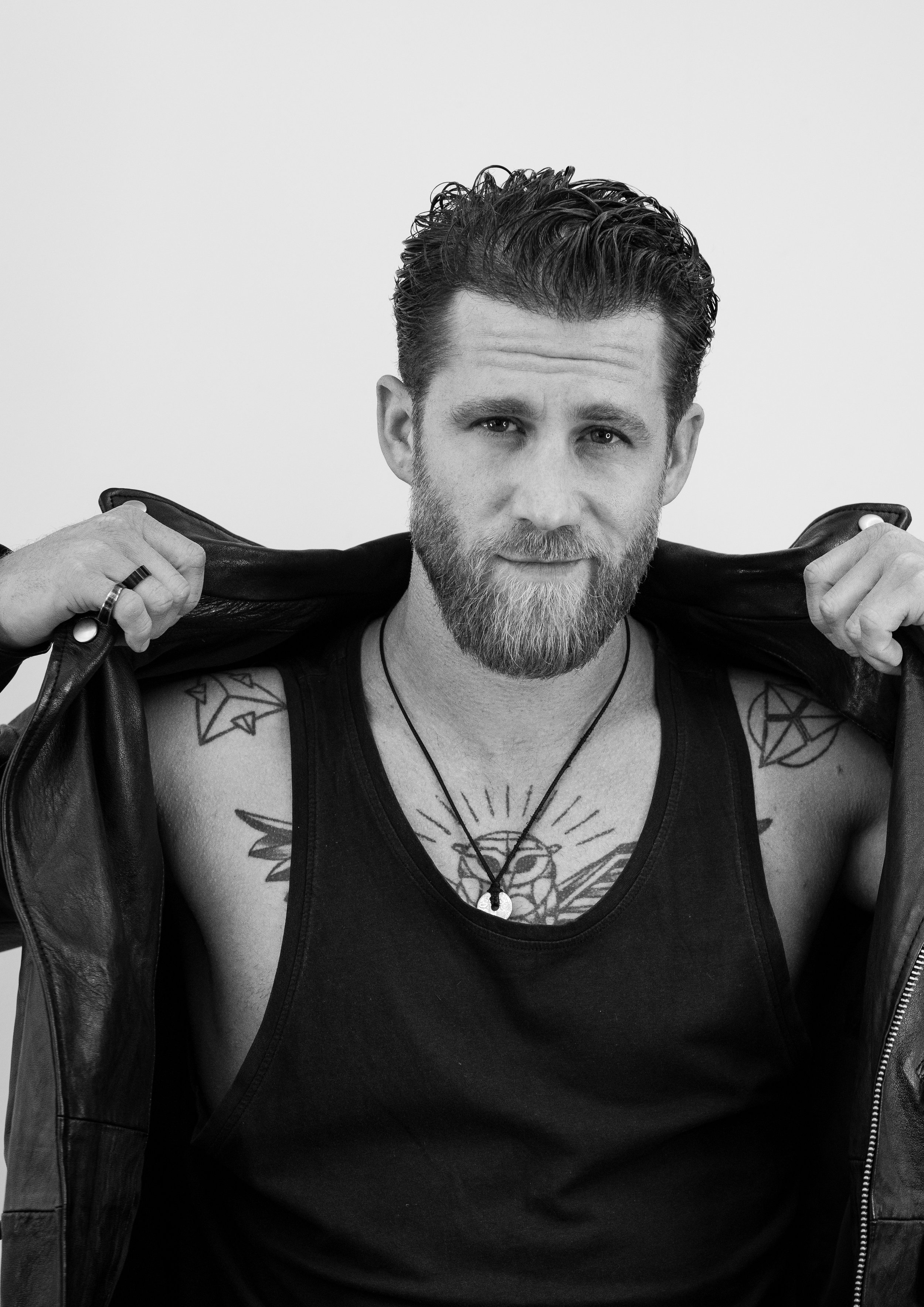 Černobílý portrét muže s plnovousem a upravenými vlasy, v tílku a s náhrdelníkem, který si přidržuje otevřenou koženou bundu přes ramena; na obou ramenou a na hrudi jsou viditelná tetování.