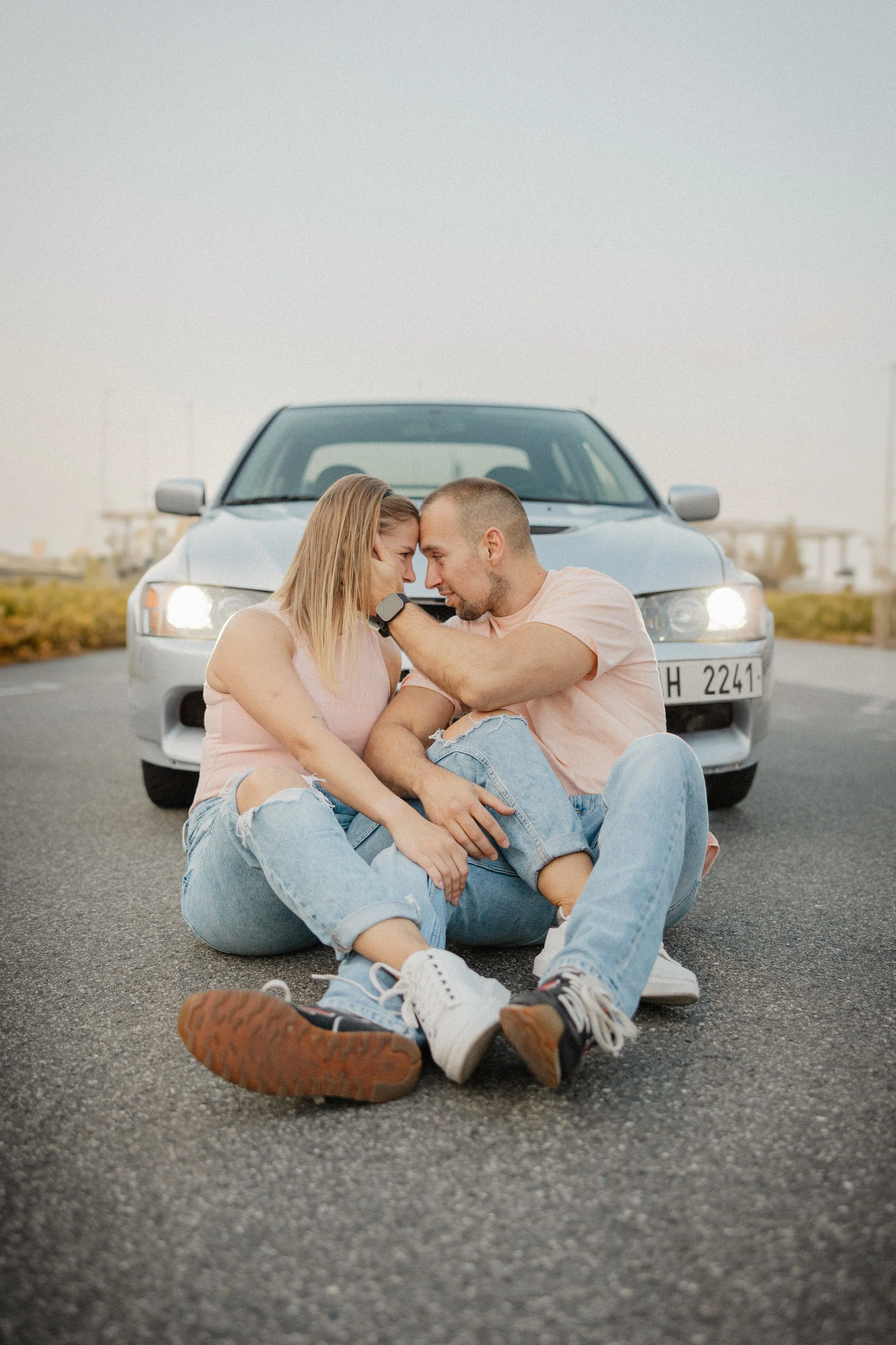 Mladý pár sedící na silnici se stříbrným autem za nimi, dotýkající se čely a usmívající se na sebe.