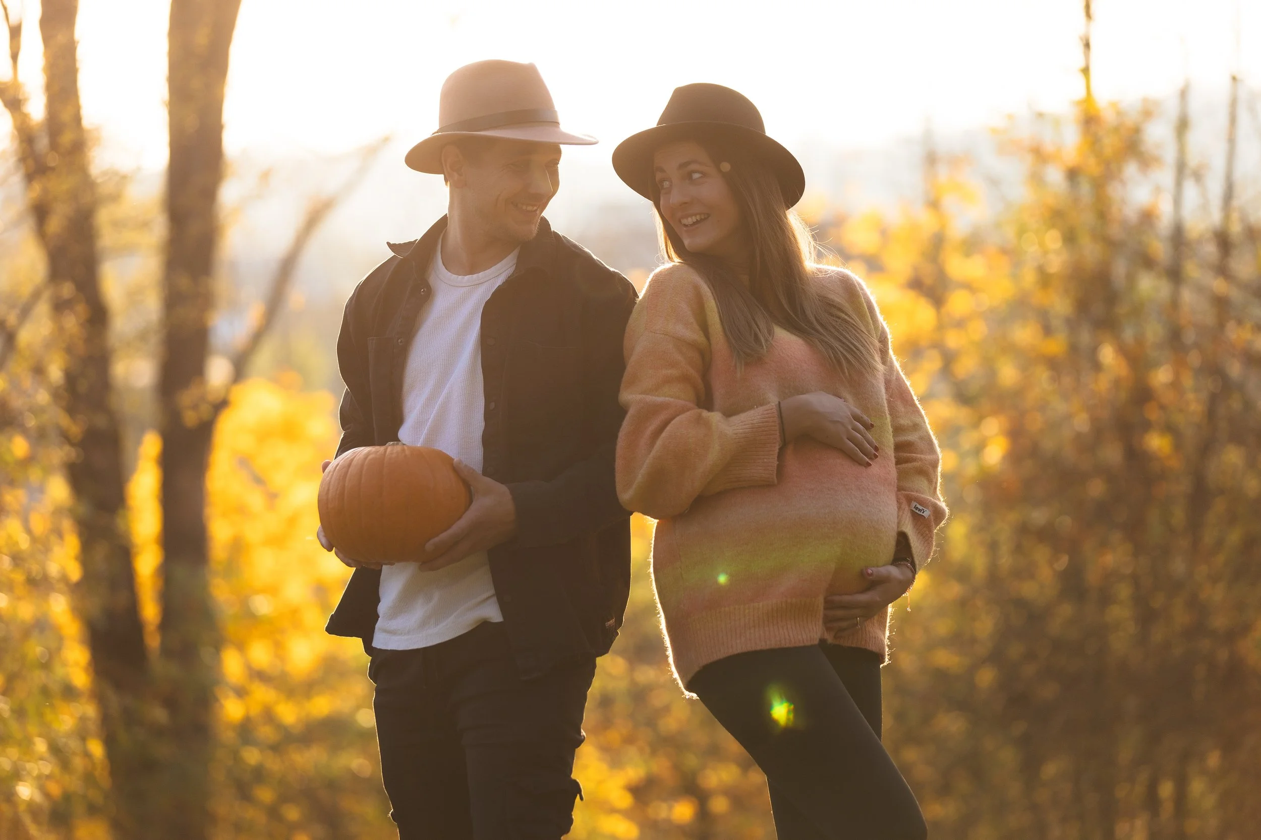 Usměvavý pár kráčí parkem na podzim, sluneční světlo prosvítá mezi stromy; muž drží dýni a žena se dotýká svého těhotenského bříška, oba mají na sobě klobouky a ležérní oblečení.