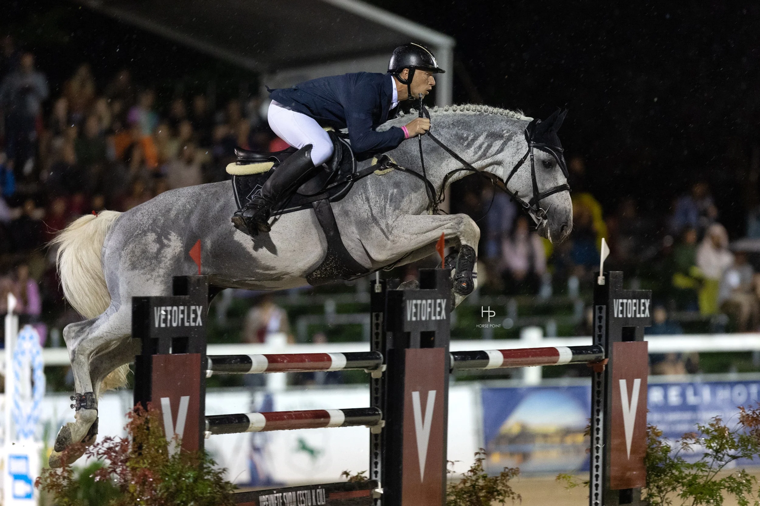 Jezdec na šedém koni ve skoku přes překážku během noční parkurové soutěže, s rozmazaným davem v pozadí.