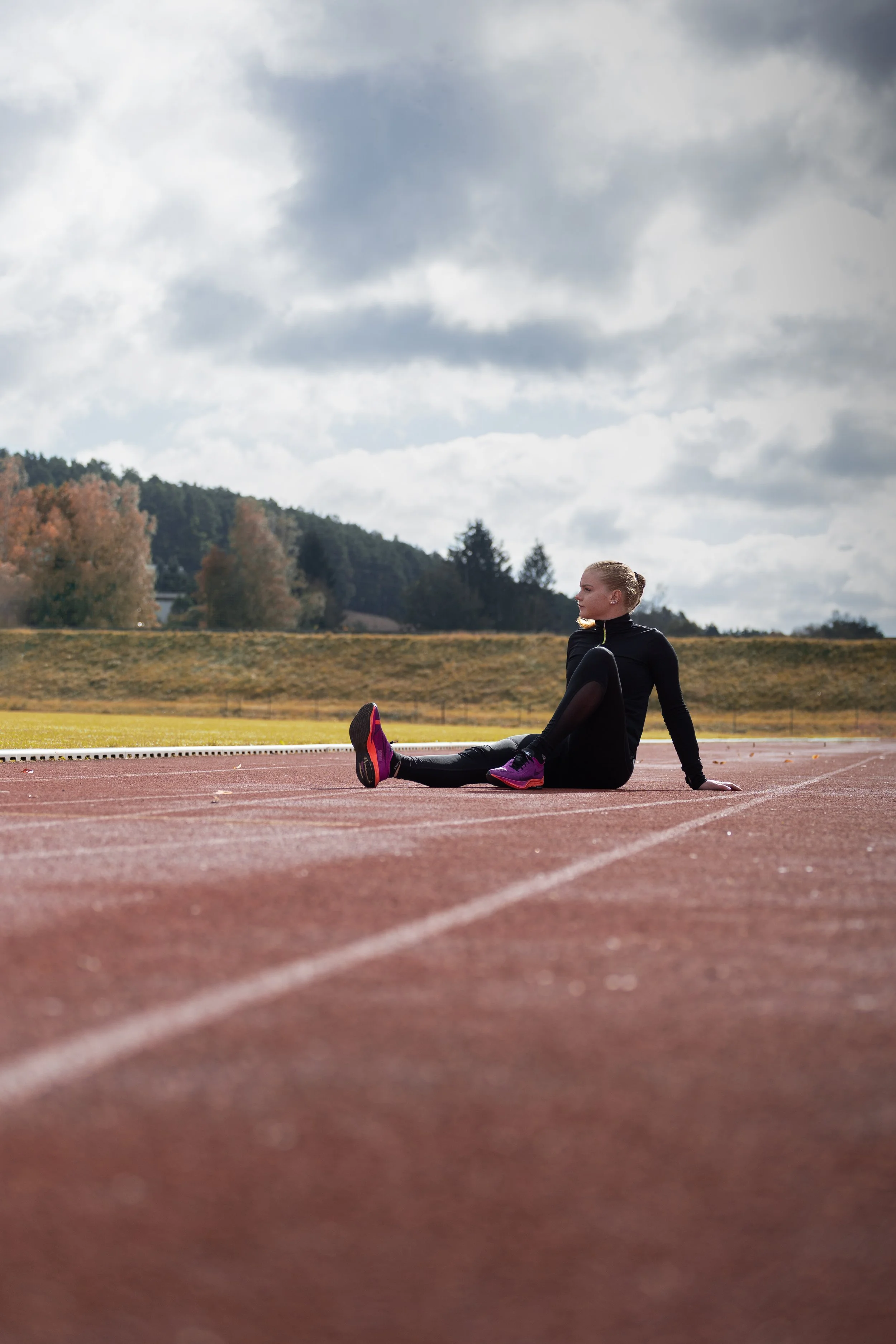 Žena ve sportovním oblečení sedí na venkovní běžecké dráze, dívá se do strany; v pozadí je zamračená obloha a stromy.