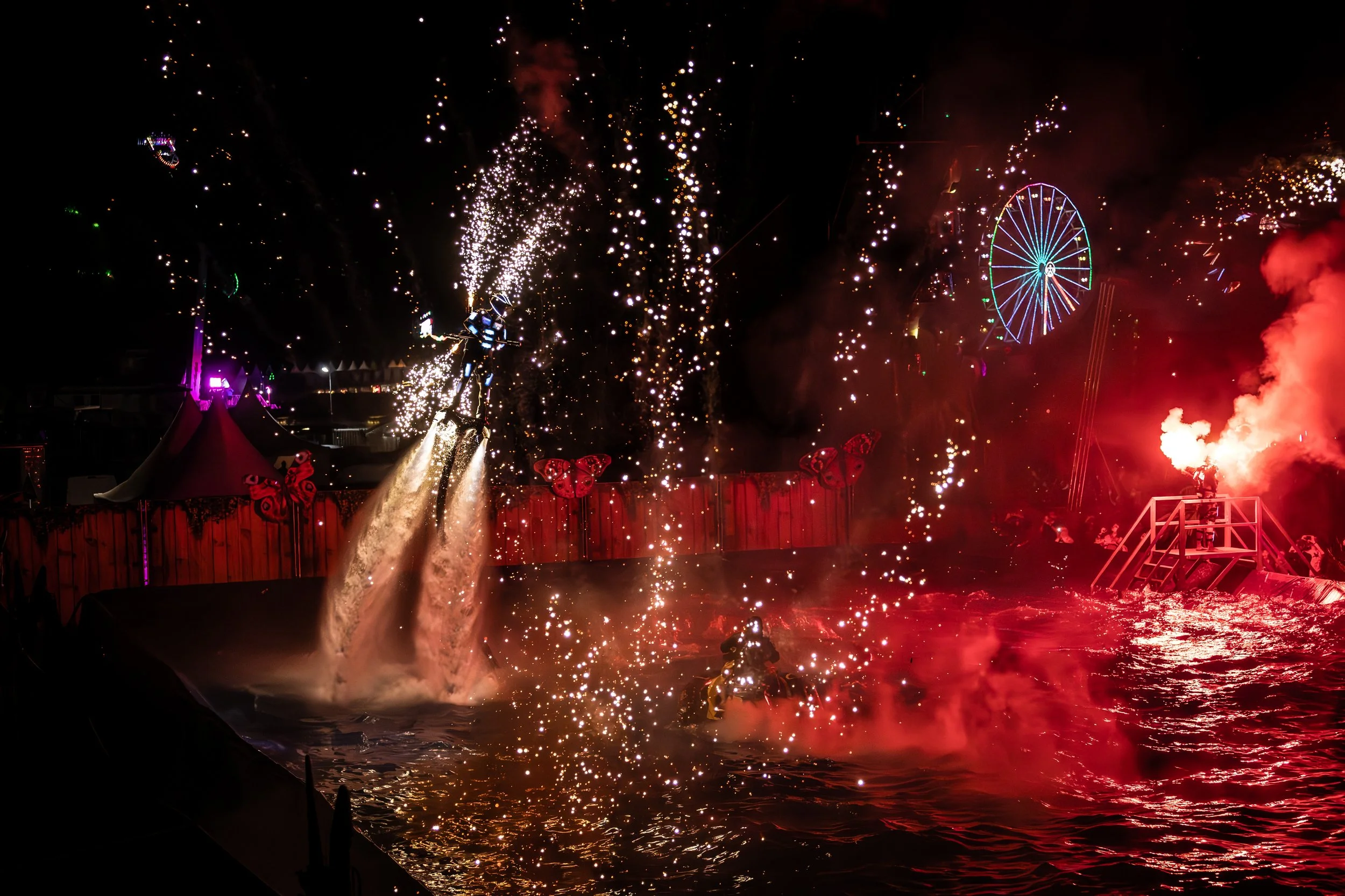 Noční scéna z lunaparku s ohňostrojem, jasnými červenými a fialovými světly, ruským kolem a vodní show s osobou na jetpacku, která stříká vodu a ohňostroj.