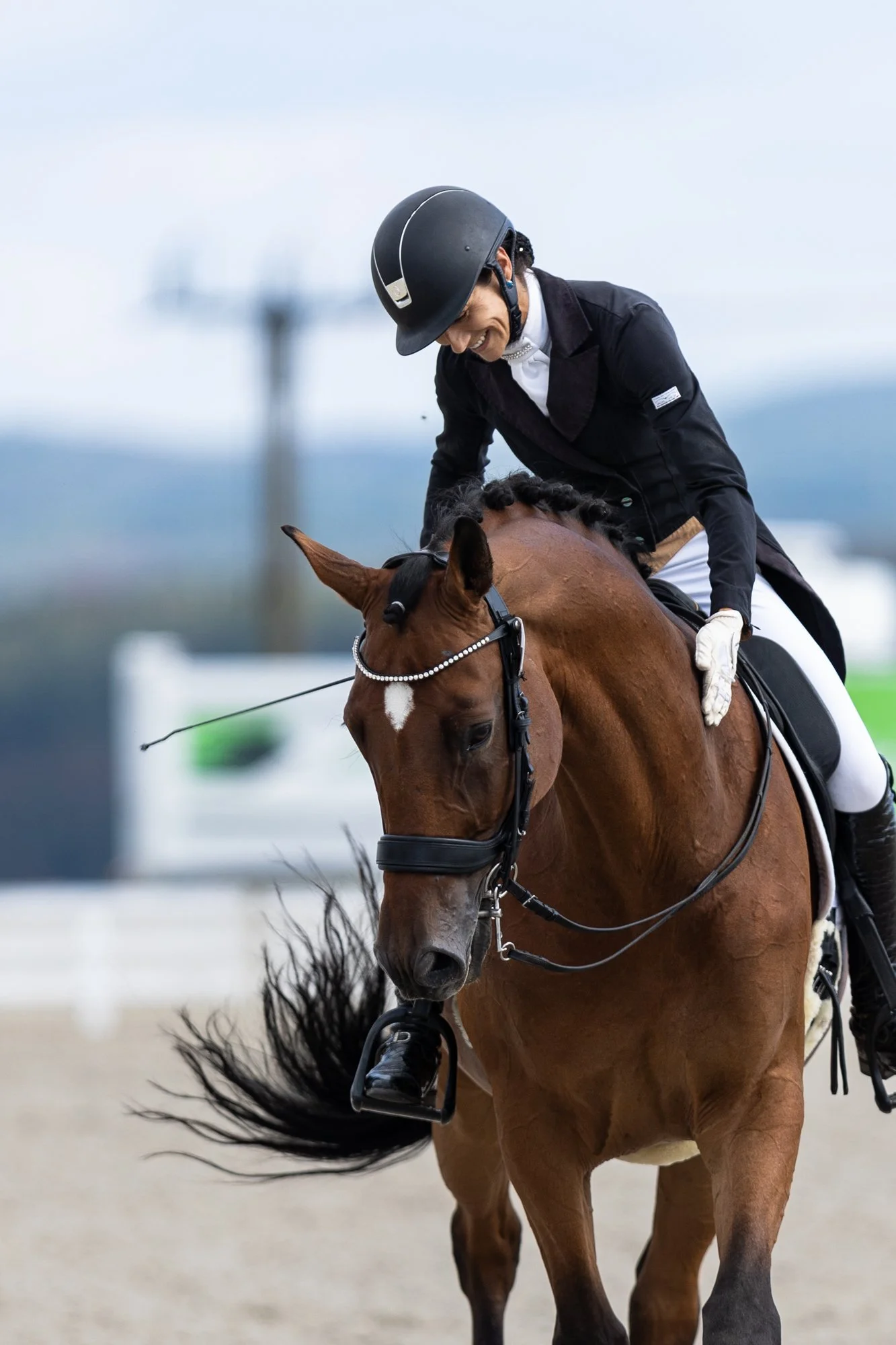 Jezdkyně se usmívá při jízdě na hnědém koni během soutěže; oba jsou v jezdeckém vybavení, s rozostřeným venkovním pozadím.
